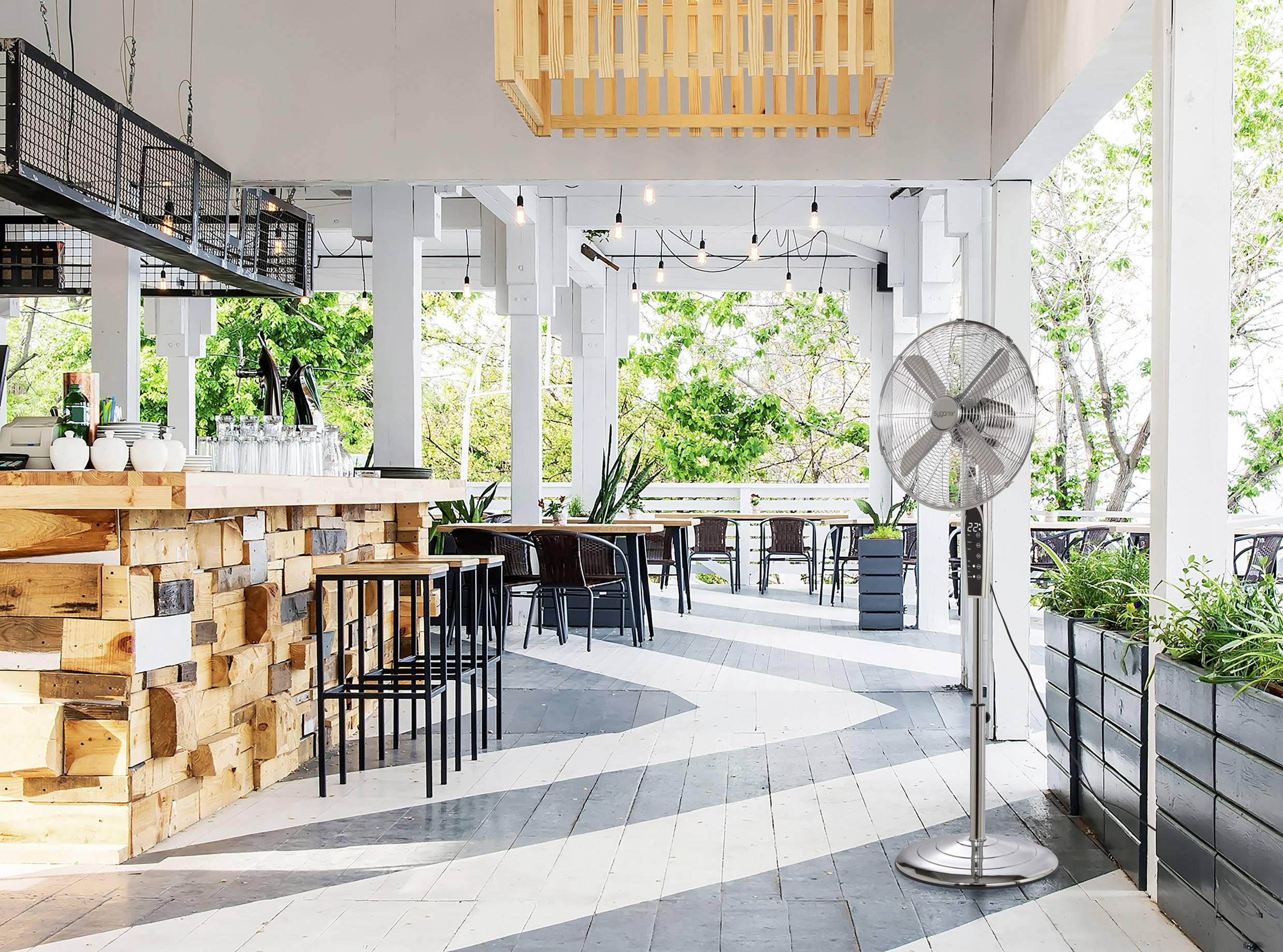 Ein Straßencafé mit Holzeinrichtung, hohen Hockern und einem Deckenventilator. Umgeben von Grünpflanzen, bietet es eine entspannte und luftige Atmosphäre.