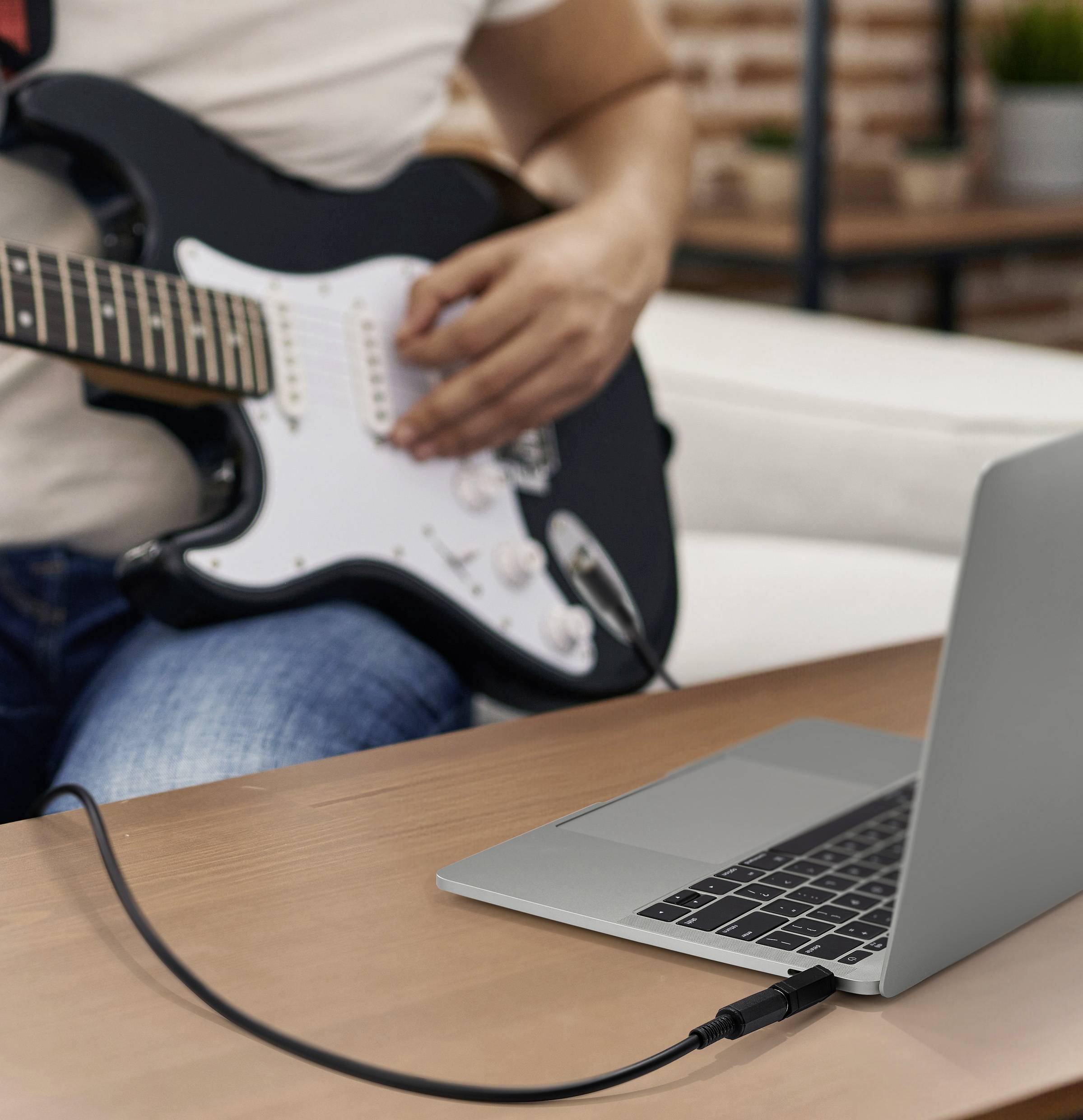 Person spielt E-Gitarre, die mit einem Kabel an einen Laptop angeschlossen ist und auf Musikaufnahme oder digitale Klangverarbeitung hindeutet.
