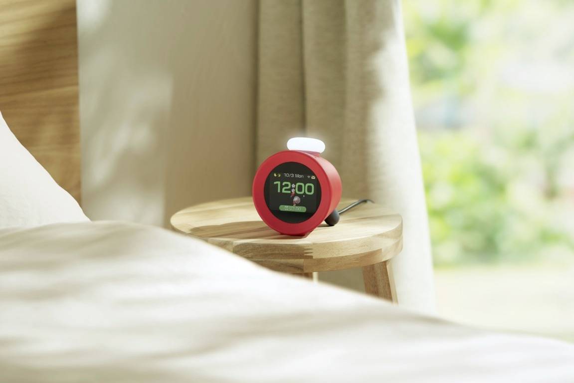 Eine Hand stellt einen roten digitalen Wecker, der '21:00' anzeigt, auf einem Holznachttisch neben einer beleuchteten Lampe ein.