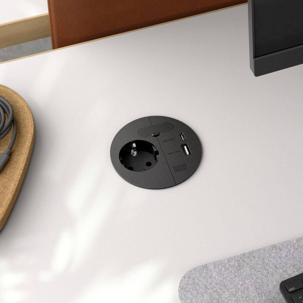 Eine Steckdose und ein USB-Anschluss, die in einen Schreibtisch eingelassen sind, mit einem Monitor, der daneben positioniert ist und einen Arbeitsplatz-Aufbau andeutet.