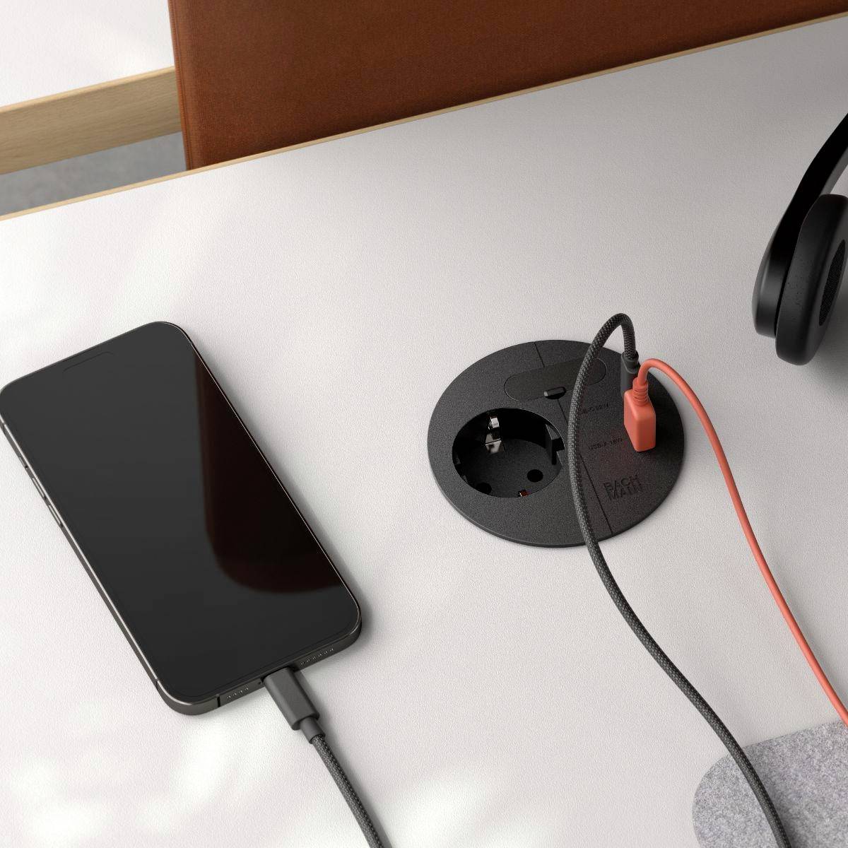 Ein Telefon und Kopfhörer auf einem Tisch, wobei das Telefon über ein Kabel, das mit einer eingebauten kreisförmigen Steckdose verbunden ist, aufgeladen wird.