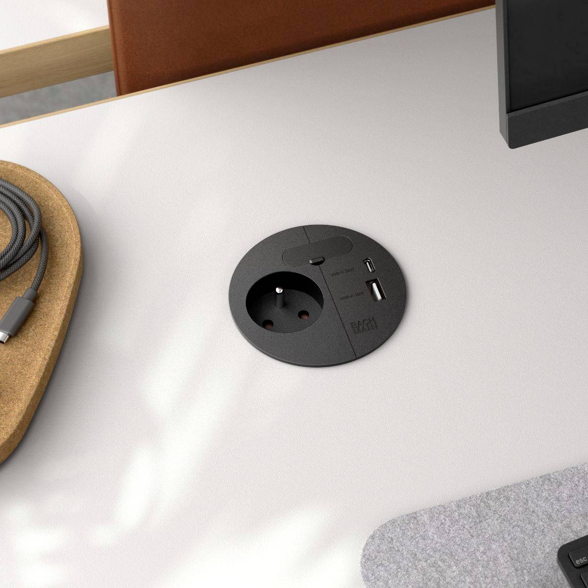 Ein runder Schreibtisch mit integriertem Stromverteiler, ausgestattet mit einer Steckdose und einem USB-Anschluss, neben einem Ledersessel und einer grauen Filzmatte in einer Büroumgebung.