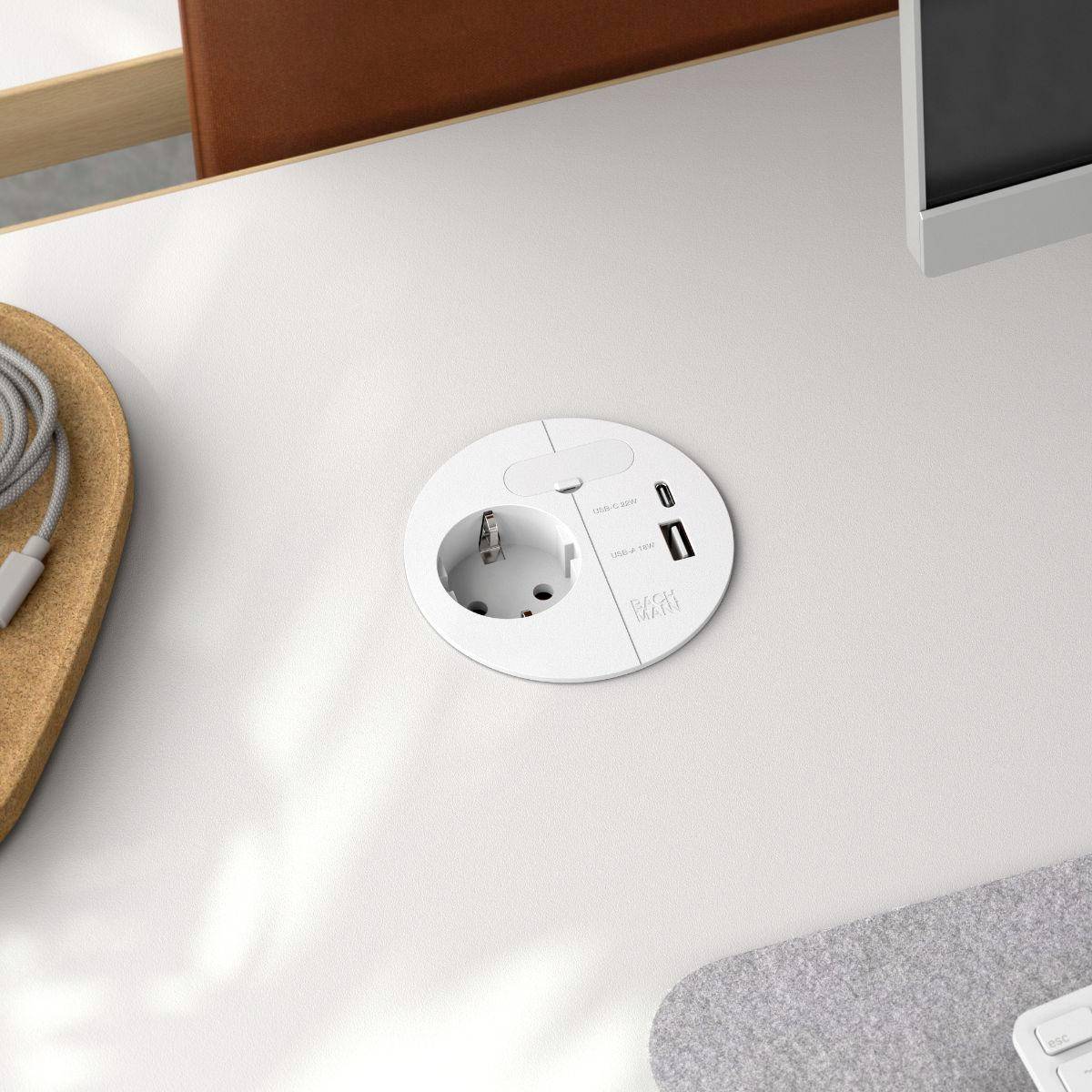 Ein weißer Schreibtisch mit integrierter Steckdose und USB-Anschluss, ein Computermonitor an der Seite und ein Kabel auf einem Holztablett.