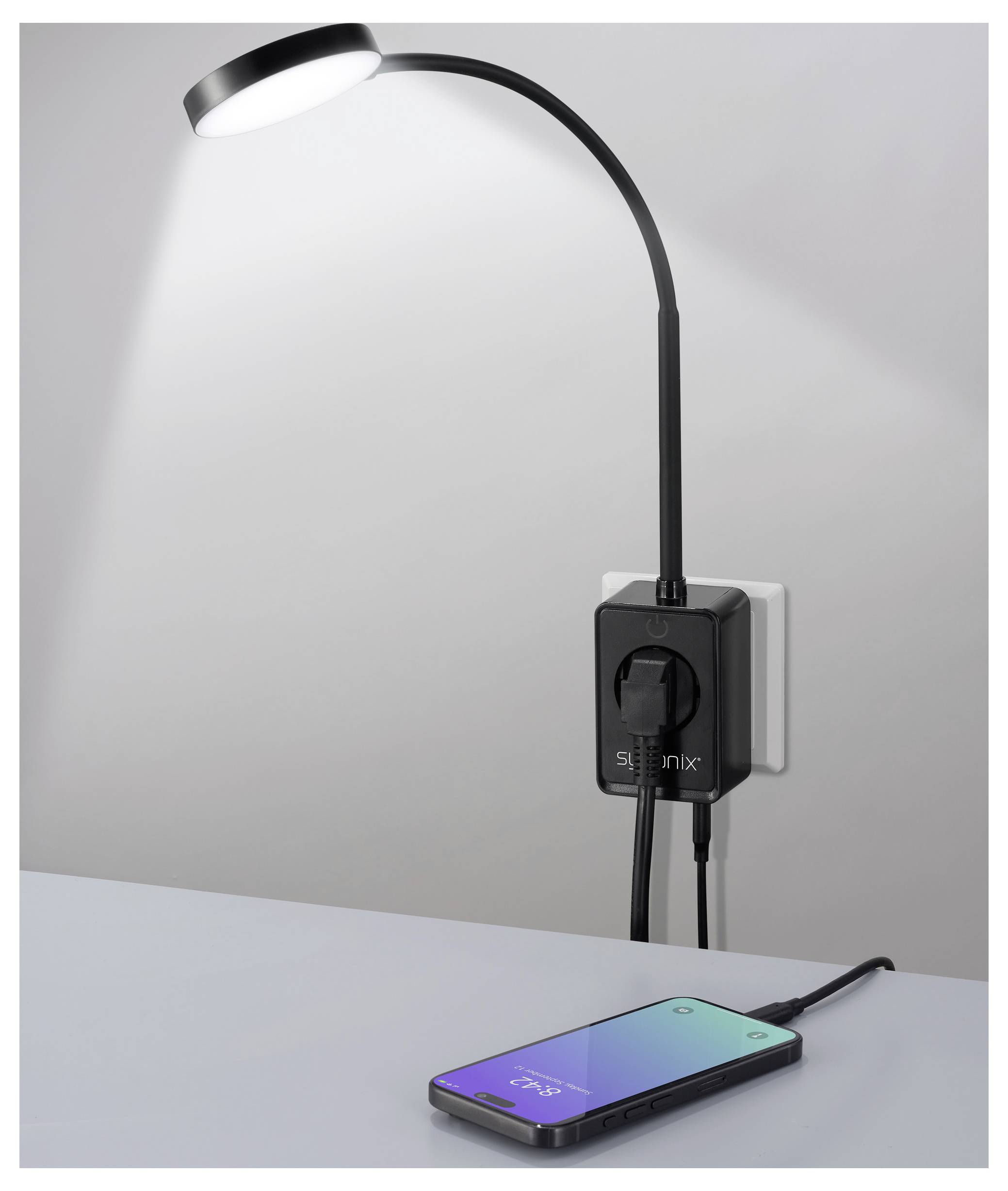 Eine flexible schwarze Lampe, die in eine Wandsteckdose eingesteckt ist, beleuchtet ein Smartphone, das auf einem Tisch liegt und einen farbigen Bildschirm zeigt.