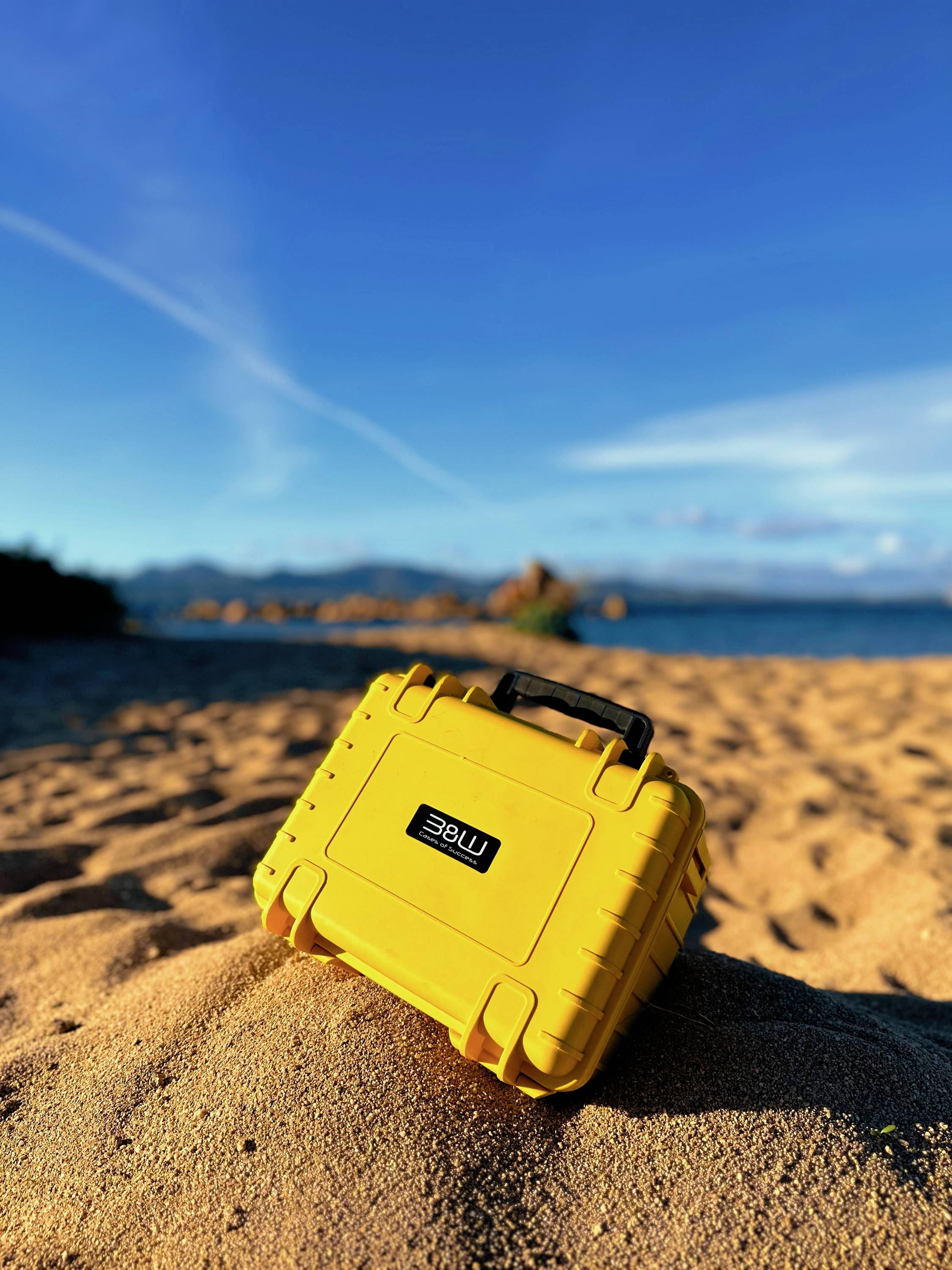 Ein hellgelbes Schutzetui liegt auf einem sandigen Strand unter einem klaren blauen Himmel, mit entfernten Bergen und dem Meer im Hintergrund.