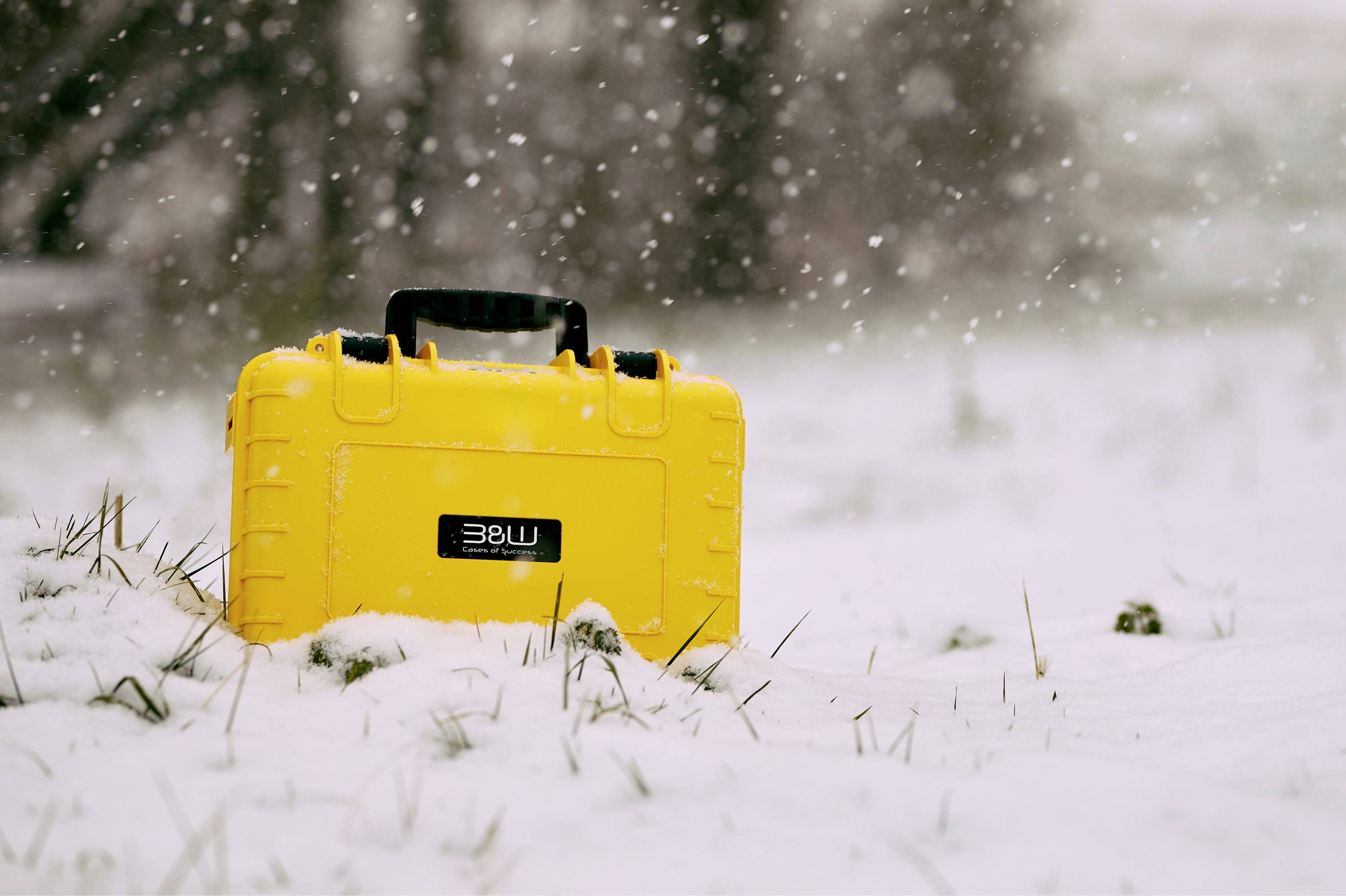 Ein heller gelber Koffer mit einem Griff steht auf verschneitem Boden, umgeben von fallenden Schneeflocken, was Outdoor-Ausrüstung oder Ausrüstungsgegenstände andeutet.
