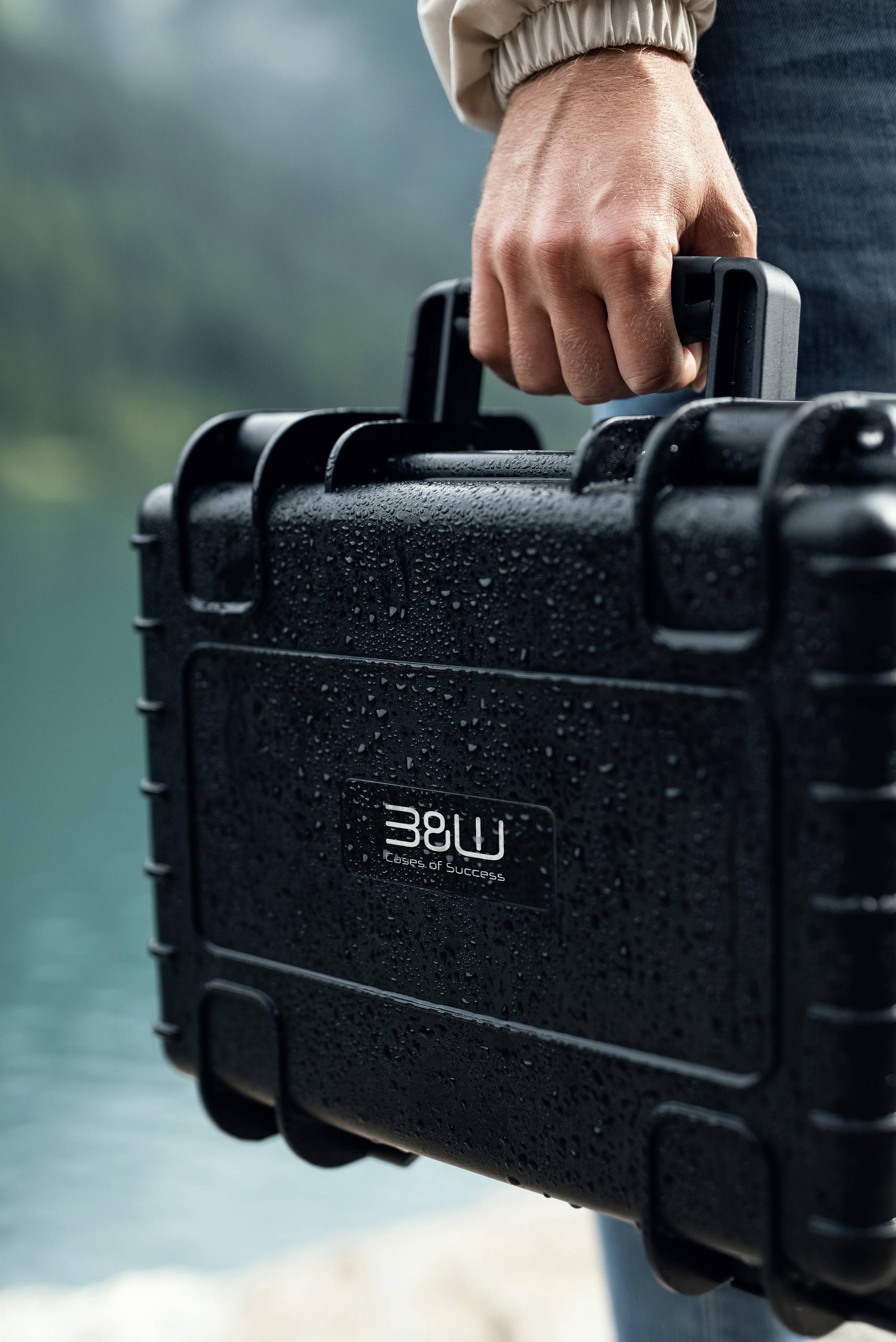 Eine Person, die einen schwarzen, wasserdichten Hartschalenkoffer an einem See hält, wobei das 'B&W'-Logo sichtbar ist. Der Koffer ist mit Regentropfen bedeckt, was auf feuchte Bedingungen hindeutet.