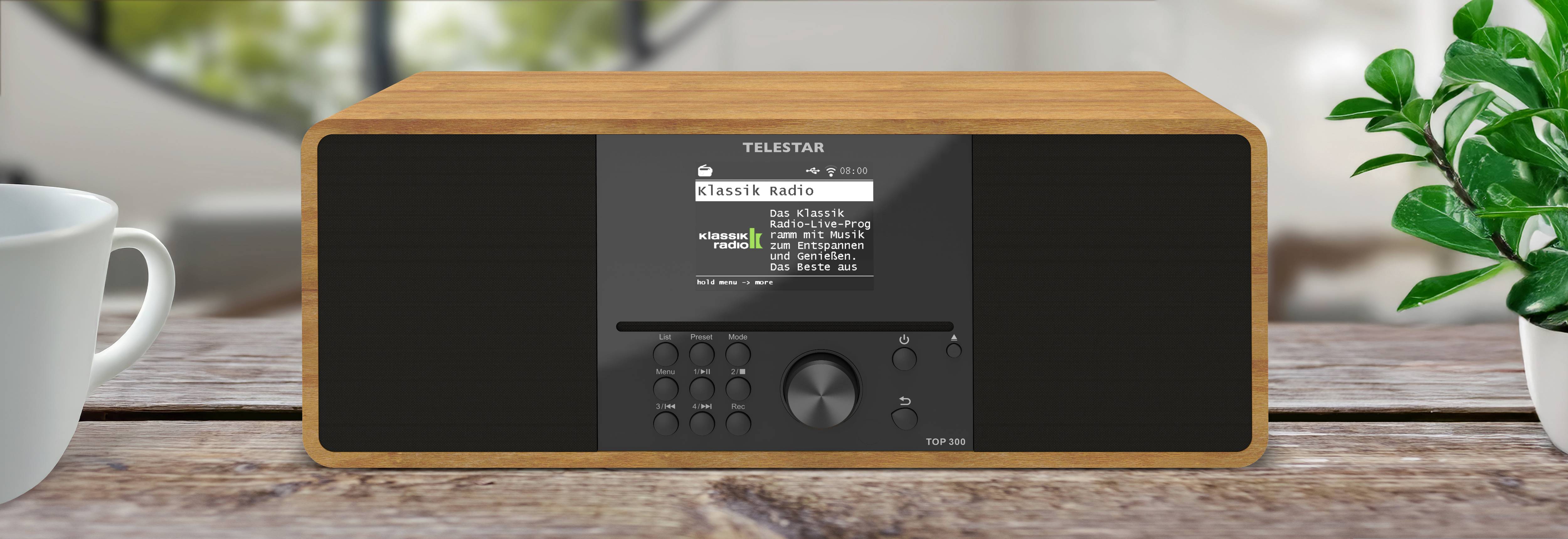 Ein rechteckiges digitales Radio mit einem Holzrahmen steht auf einem Holztisch und verfügt über einen Displaybildschirm, Bedienungsknöpfe und einen Drehregler.