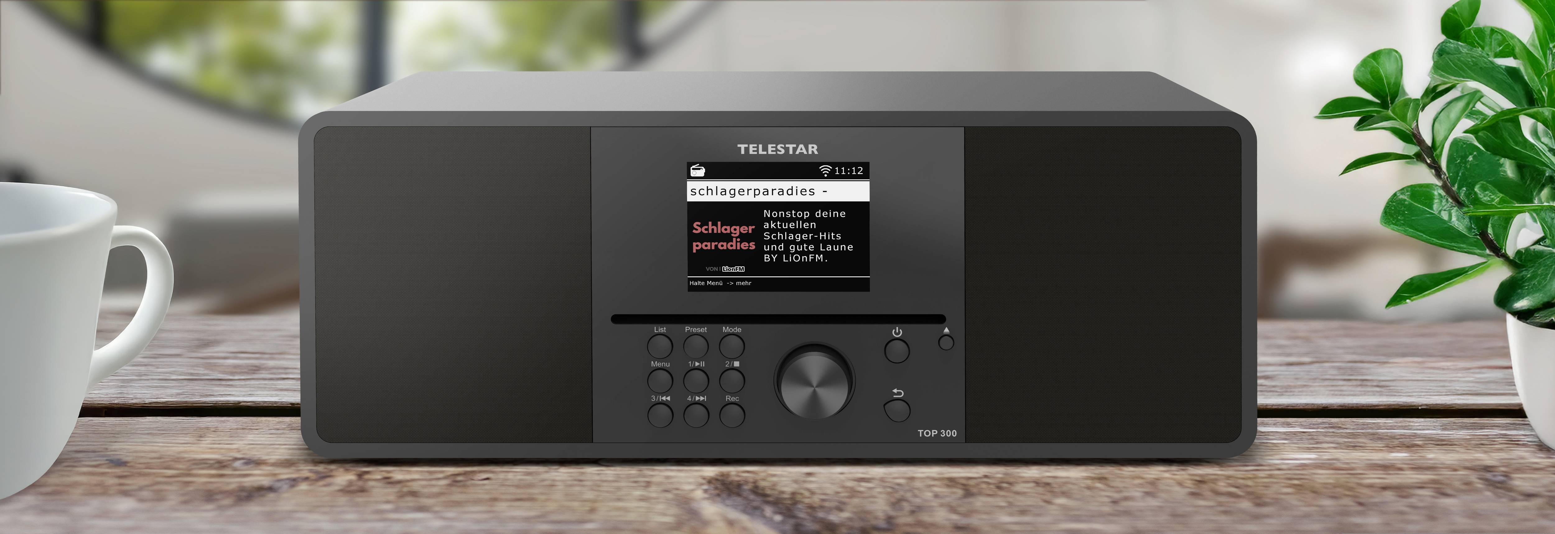 Ein schwarzes TELESAR Digitalradio steht auf einem Holztisch mit einer Anzeige, die ‚Schlagerparadies' zeigt, umgeben von einer Tasse und einem Fensterblick.