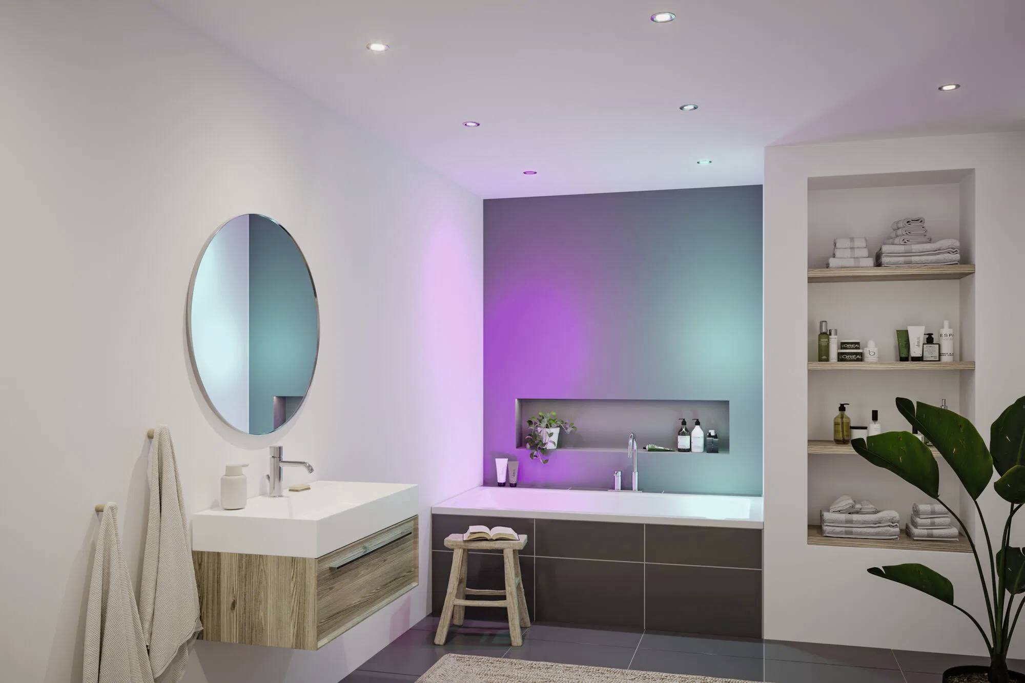 Zeitgenössisches Badezimmer mit einem runden Spiegel, schwebendem Waschbecken, Badewanne und Stimmungsbeleuchtung. Regale mit Handtüchern; eine Pflanze bringt Grün.