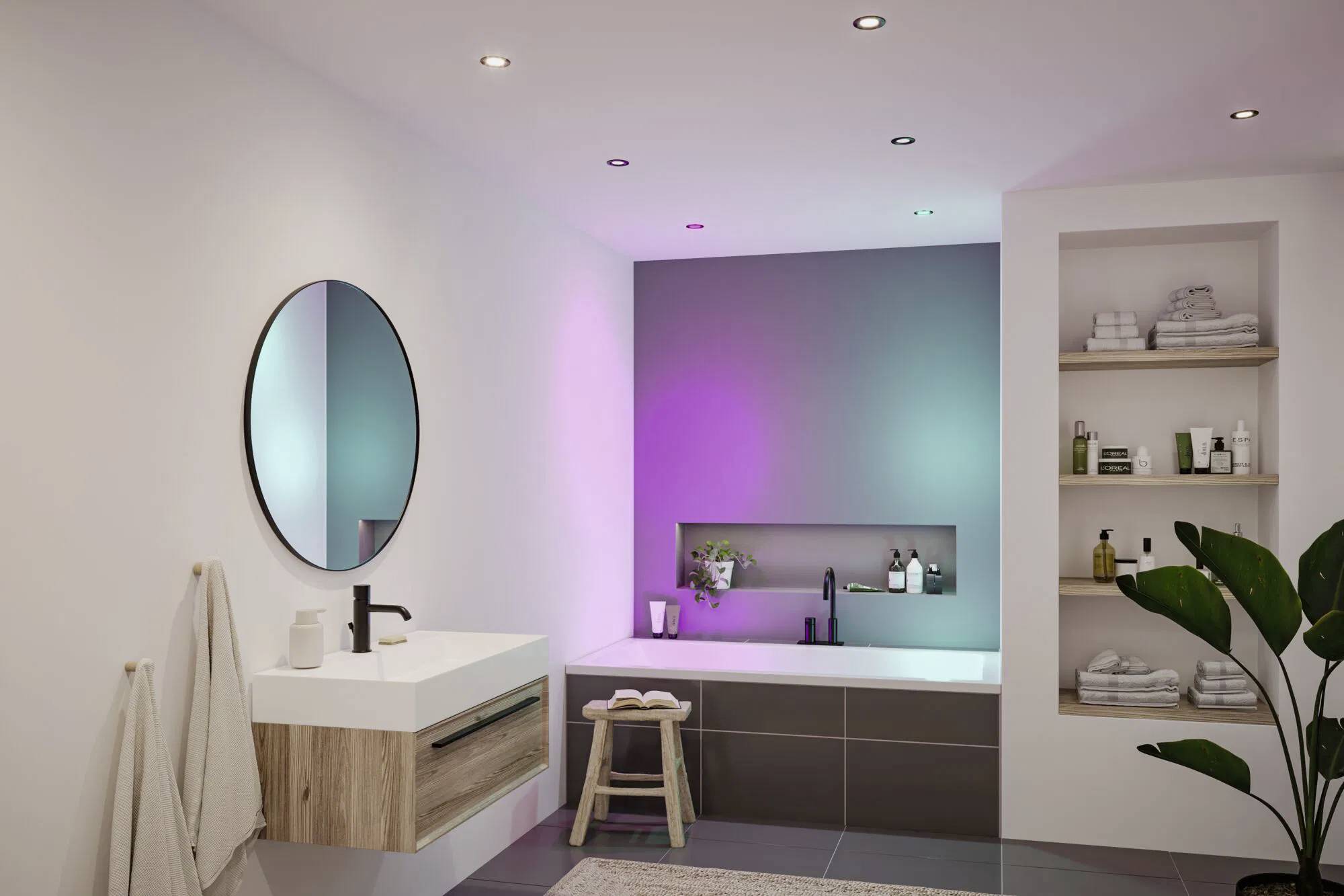 Ein modernes Badezimmer mit einem runden Spiegel, einem wandmontiertem Waschbecken, einer Badewanne unter bunten Lichtern und einem Regal mit Handtüchern und Toilettenartikeln.