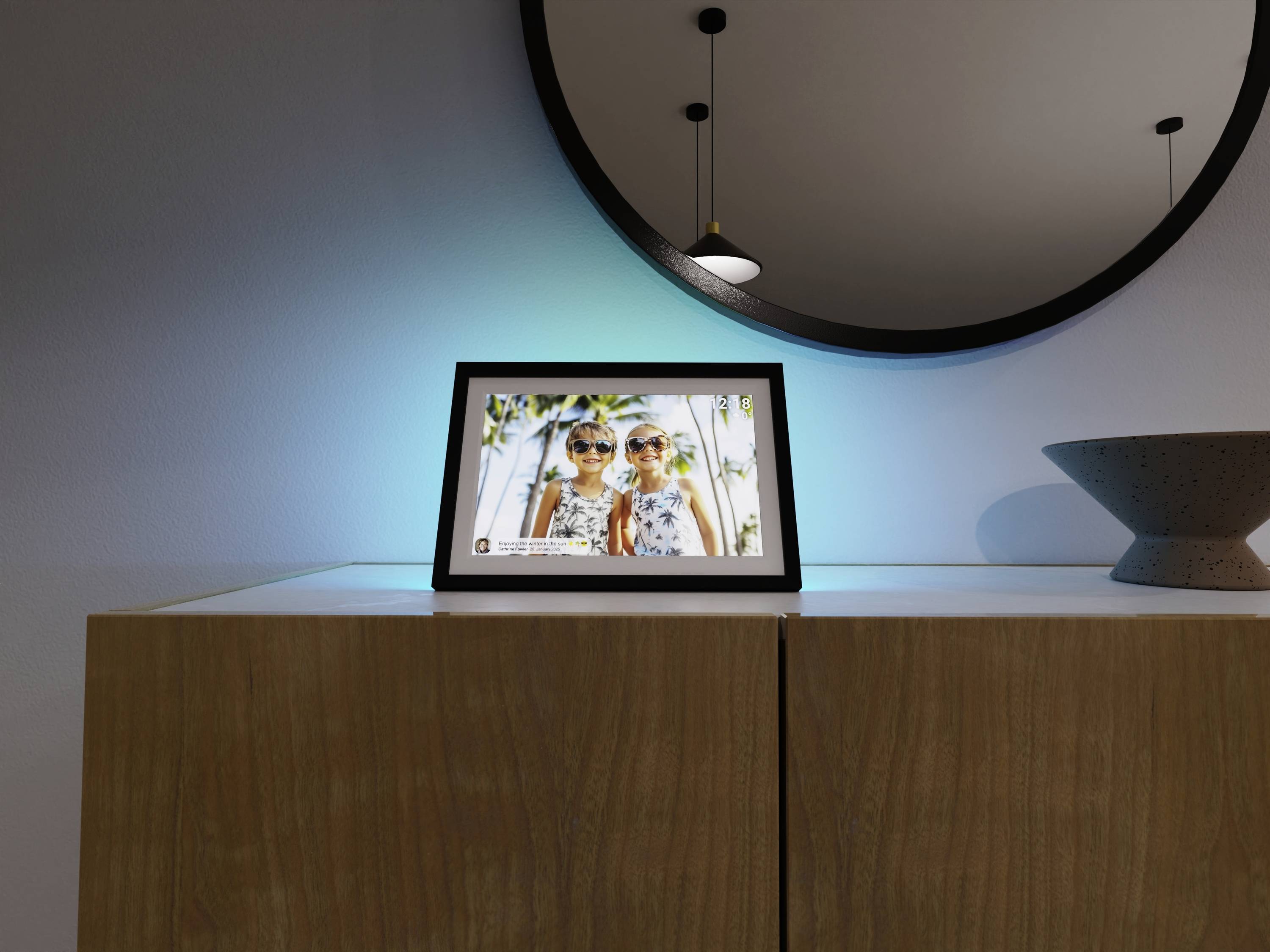 Ein digitaler Fotorahmen auf einem Holzschrank unter einem runden Spiegel zeigt ein Bild von zwei lächelnden Kindern, mit einer modernen Leuchte darüber.