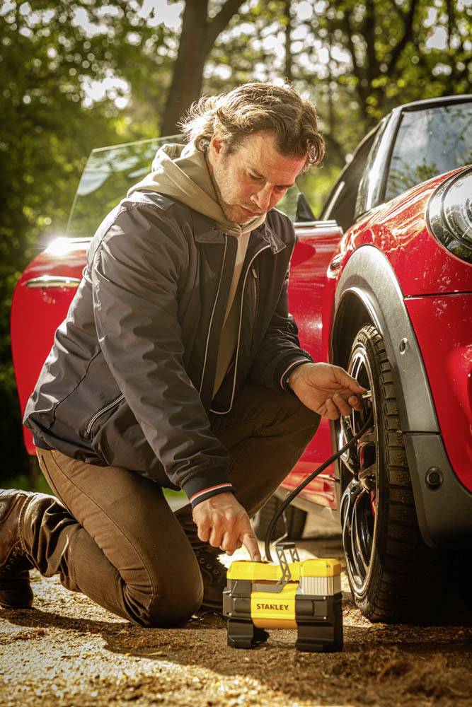 Mann kniet neben einem roten Auto und verwendet einen tragbaren Luftkompressor, um einen Reifen aufzupumpen, umgeben von einer Waldlandschaft.