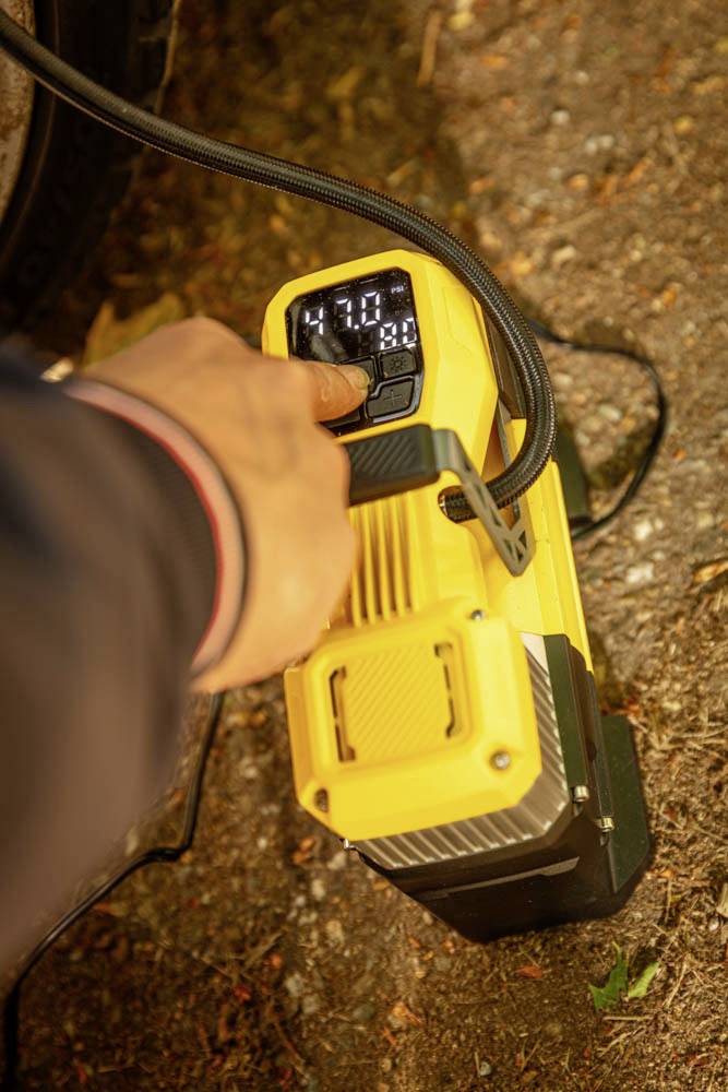 Eine Person, die das digitale Display eines tragbaren Luftkompressors einstellt, das '4,70' zeigt, aufgestellt auf einem Erdgrund neben einem Fahrzeugreifen.