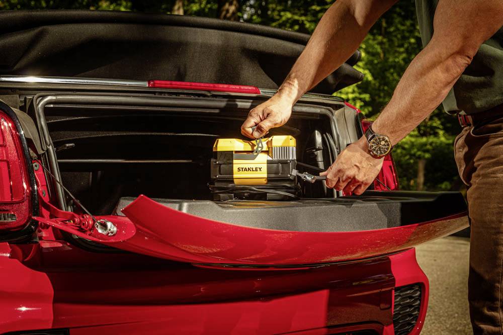 Eine Person legt eine gelbe Werkzeugkiste in den Kofferraum eines roten Autos, wobei ein Waldgebiet im Hintergrund zu sehen ist.