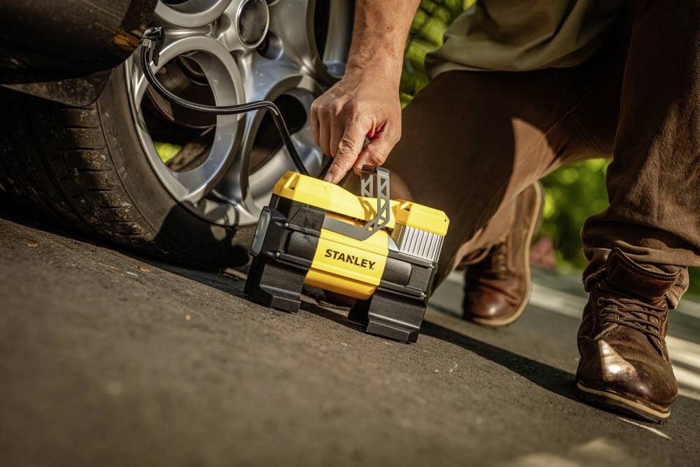 Eine Person verwendet einen gelben tragbaren Luftkompressor, um einen Autoreifen an einer sonnigen Straße aufzupumpen.