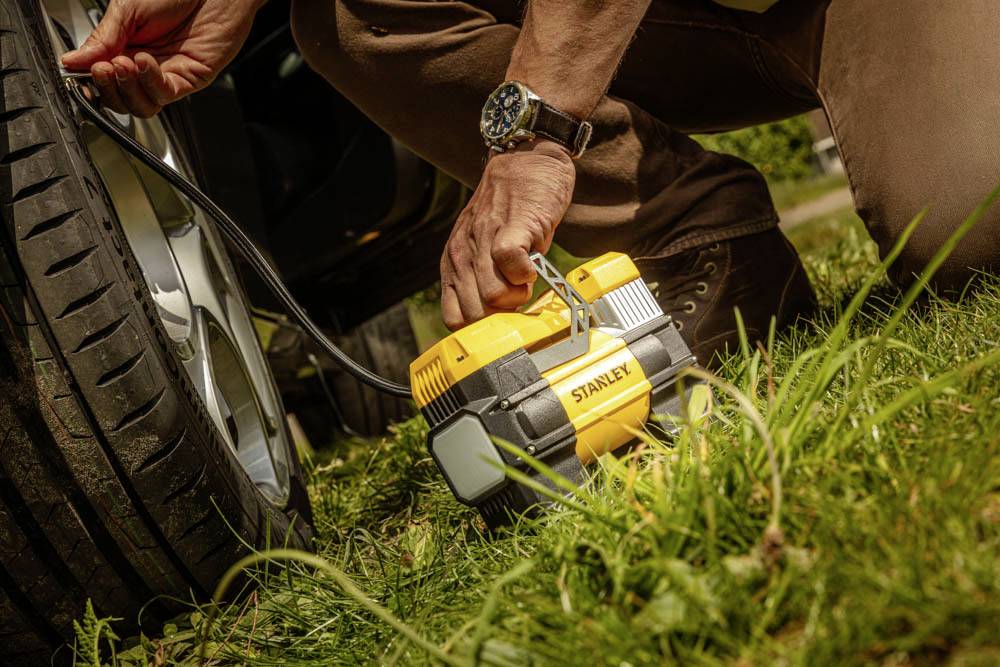 Eine Person benutzt einen tragbaren Luftkompressor, um einen Autoreifen aufzupumpen, wobei sie auf Gras kniet und das Gerät mit der Aufschrift 