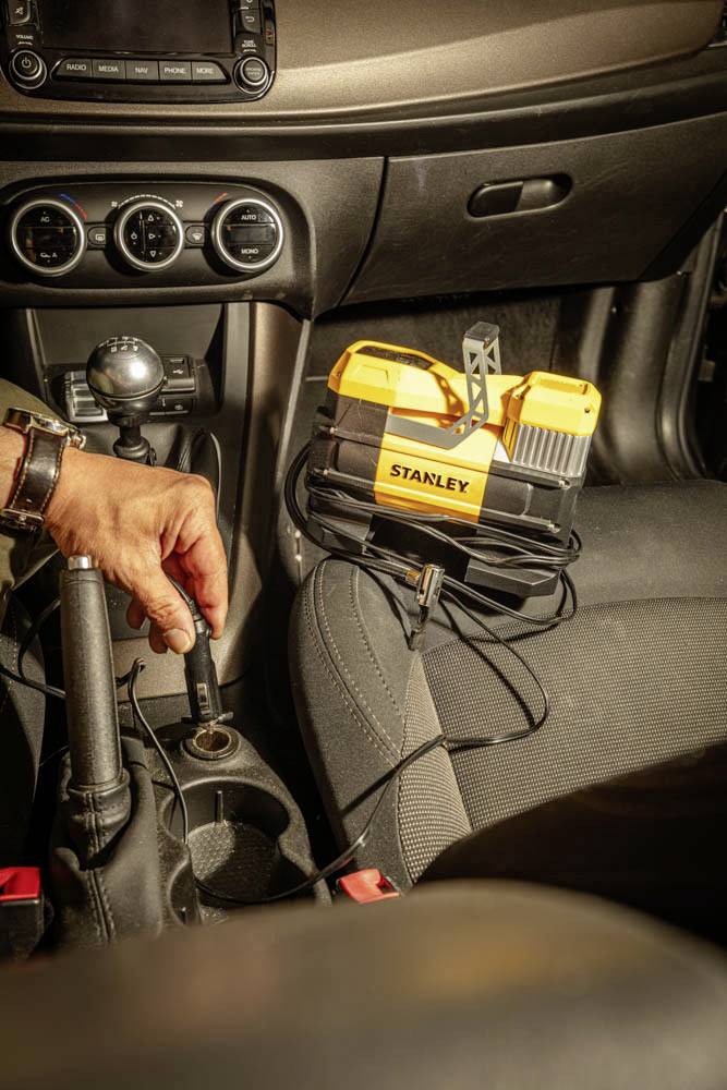 Eine Person, die einen Starthilfe-Akku in einem Auto benutzt. Das Gerät, mit dem Schriftzug 'Stanley', liegt auf dem Beifahrersitz, und eine Hand steckt es in die Stromsteckdose in der Nähe des Schalthebels.