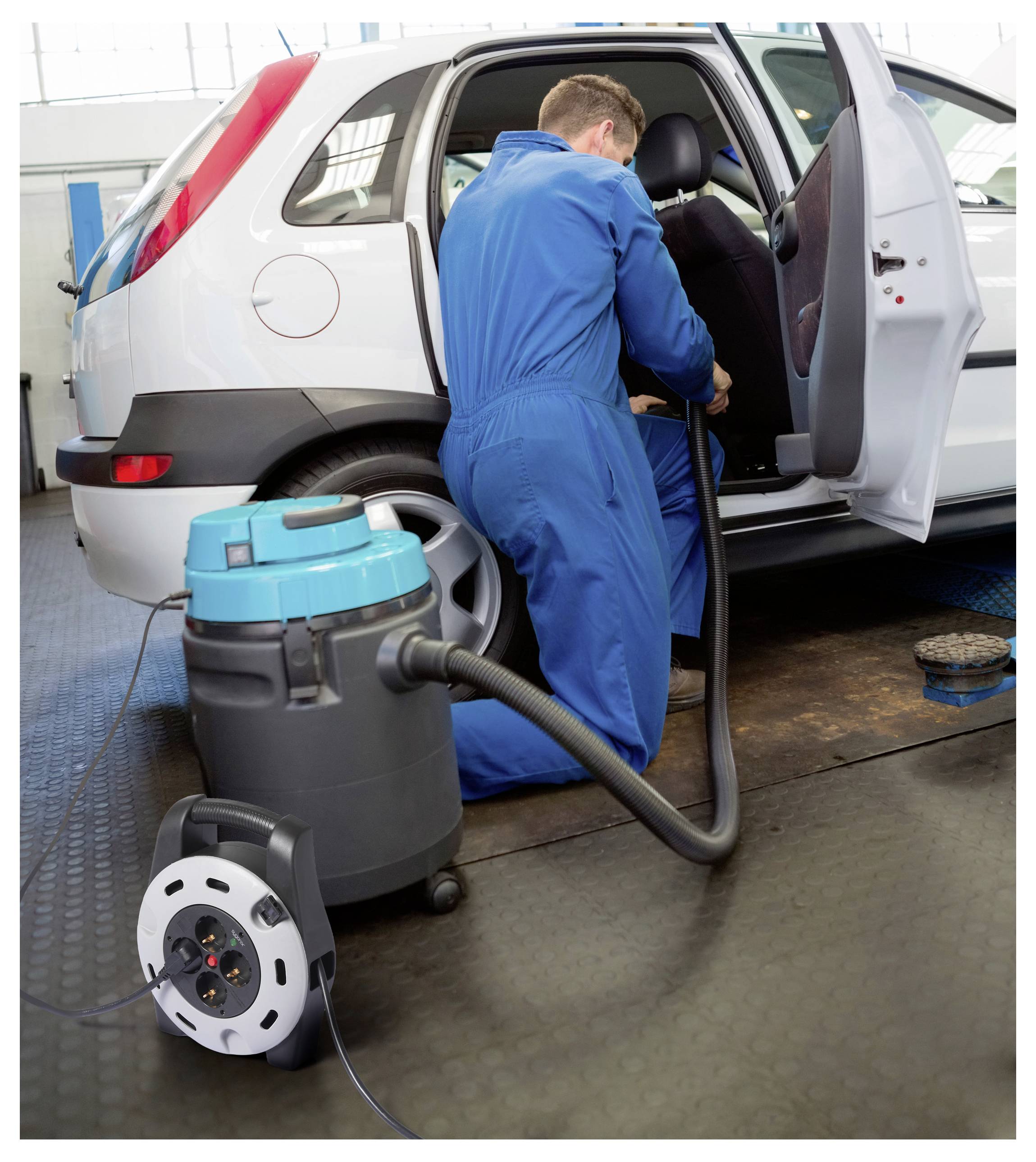 Ein Arbeiter in blauem Schutzanzug reinigt das Innere eines weißen Autos in einer Garage mit einem Staubsauger. Ein tragbarer Staubsauger steht auf dem Boden.