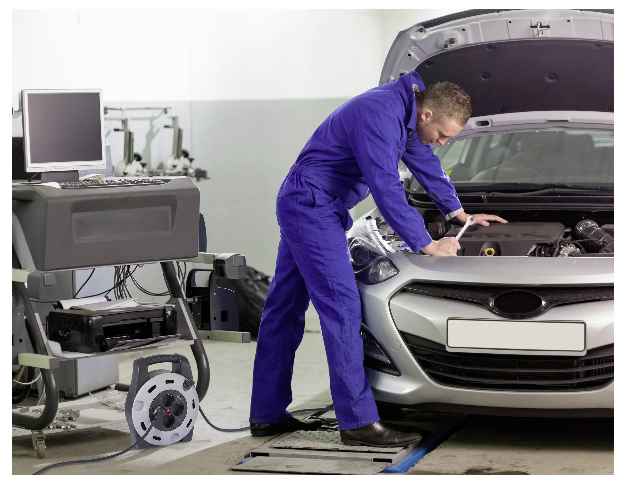 Ein Mechaniker in einem blauen Overall überprüft einen Automotor in einer Werkstatt und nutzt dabei Diagnosegeräte, die mit einem nahegelegenen Computer verbunden sind.