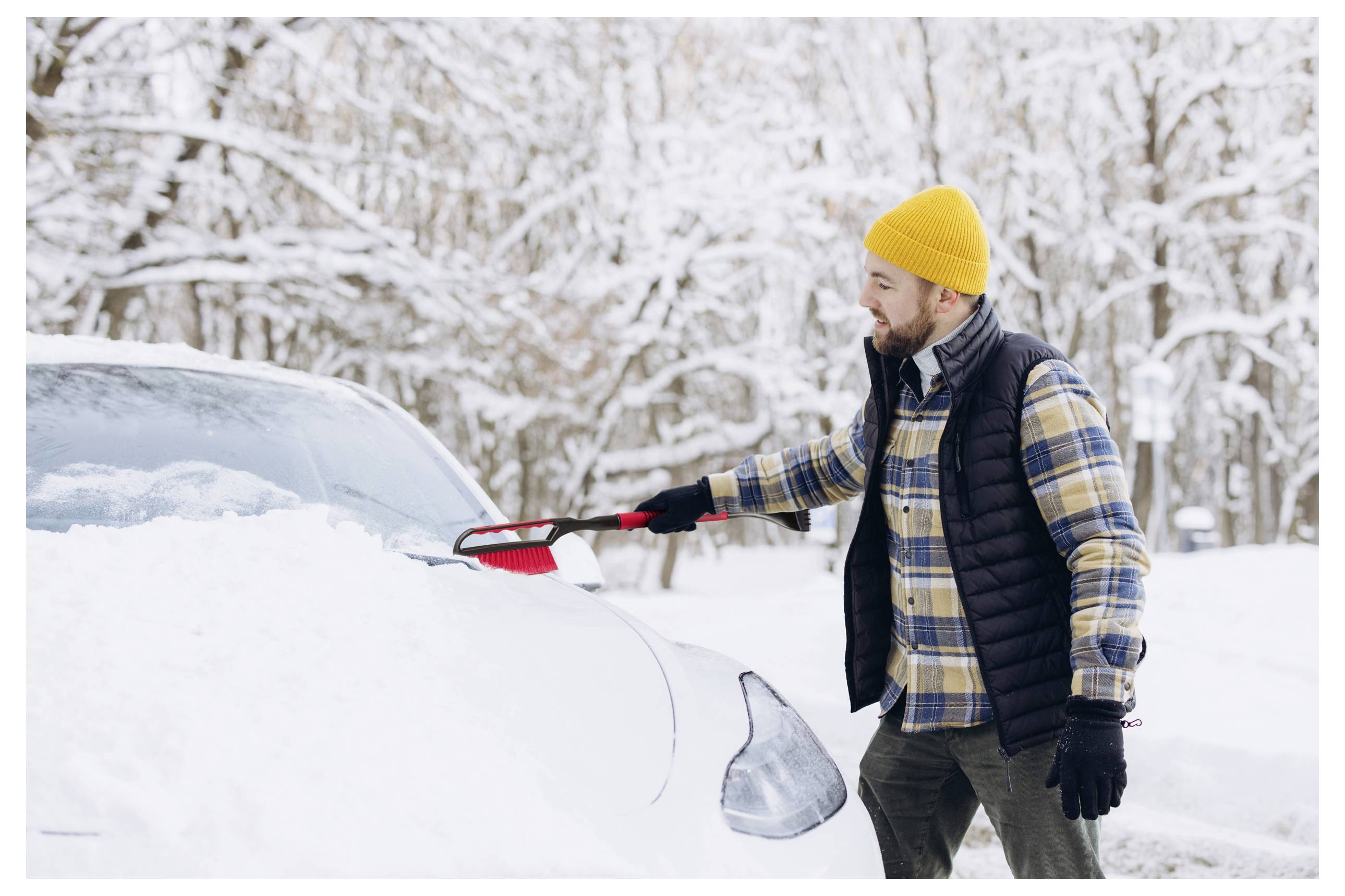 Eine Person in einem gelben Hut und einer karierten Jacke benutzt eine Schneebürste, um schweren Schnee von einem Auto in einer verschneiten Waldumgebung zu entfernen.