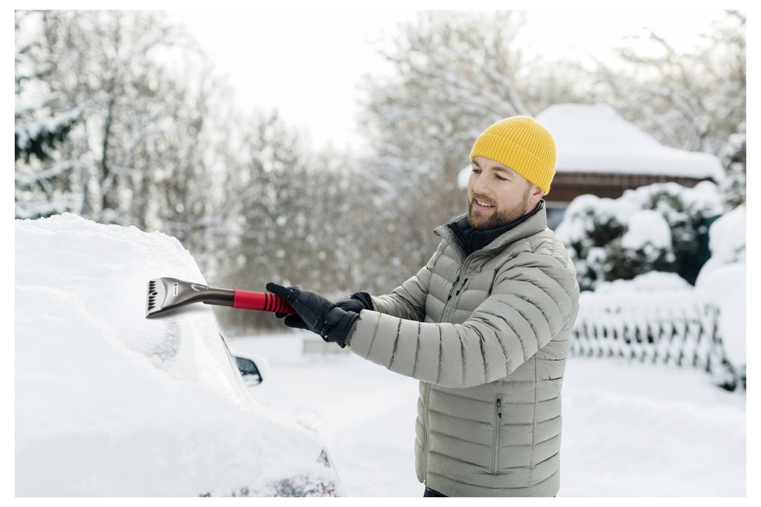 Ein Mann mit einer gelben Mütze und einer grauen Jacke räumt mit einem Eiskratzer Schnee von einem Auto in einer verschneiten Wohnumgebung.