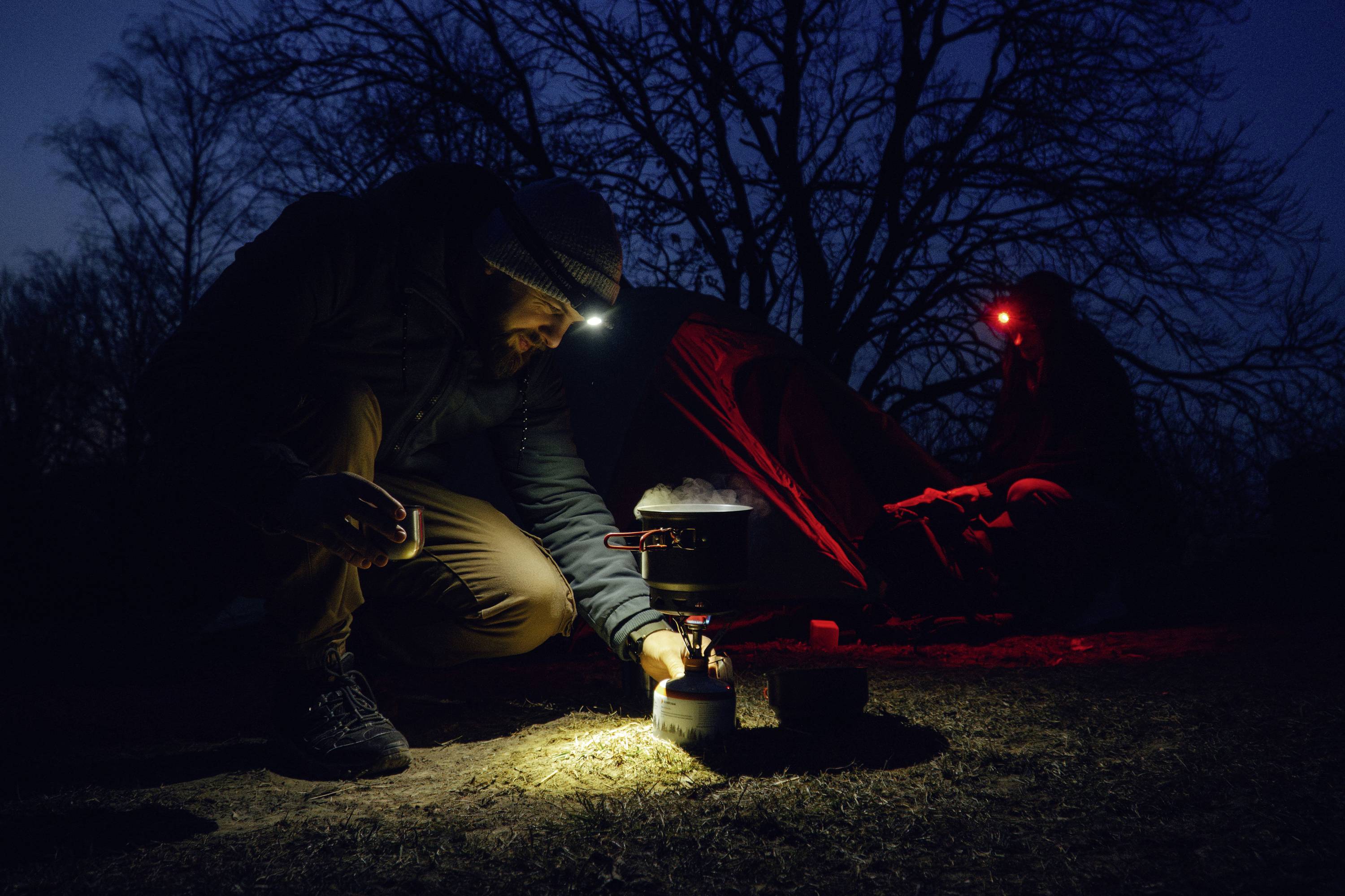 Eine Person kocht auf einem tragbaren Campingkocher neben einem Zelt bei Nacht und trägt eine Stirnlampe; eine weitere Person sitzt in der Nähe unter rotem Licht.