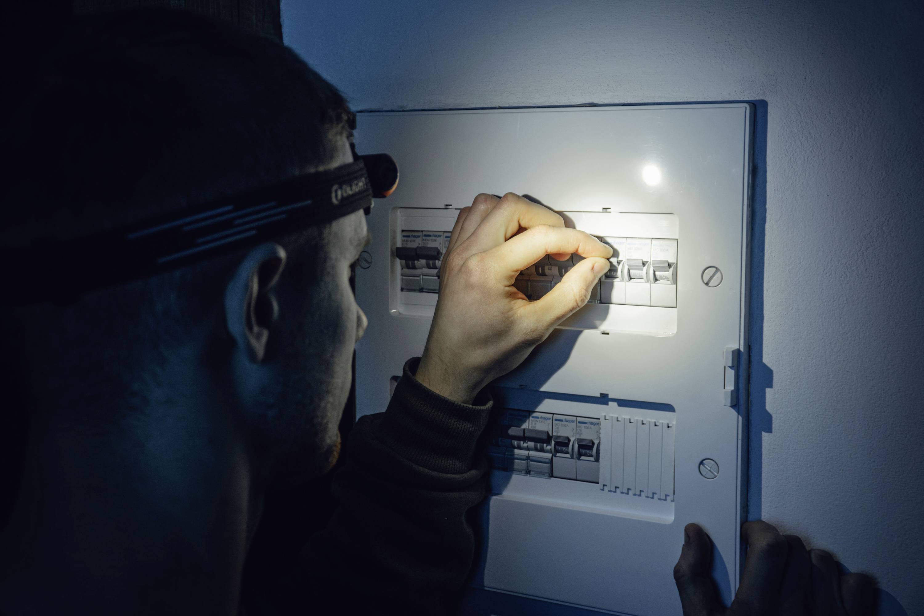 Eine Person mit einer Stirnlampe inspiziert eine elektrische Sicherungskasten und konzentriert sich darauf, einen der Schalter in einer düsteren Umgebung einzustellen.