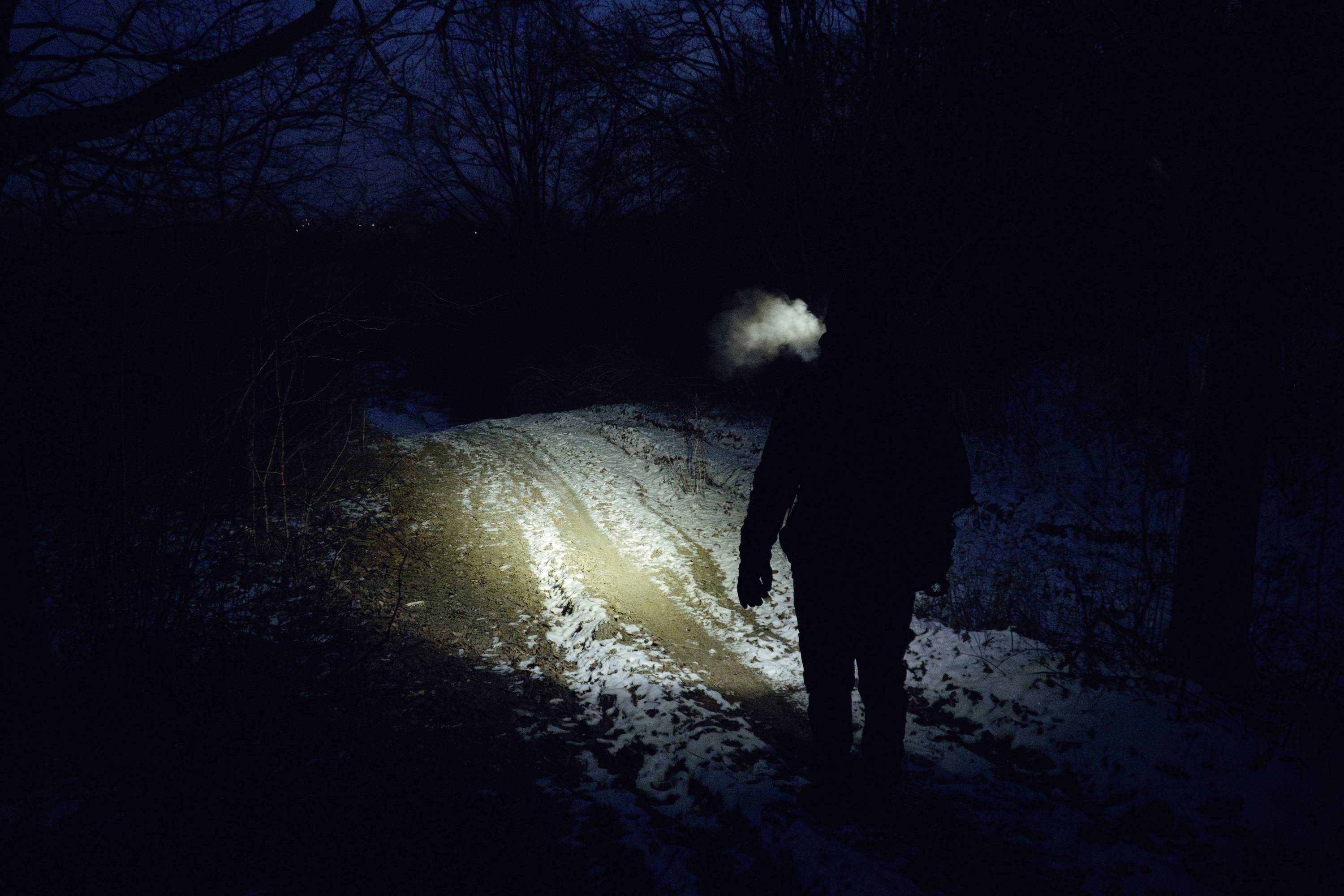 Eine Person geht einen schwach beleuchteten, verschneiten Pfad durch einen Wald bei Nacht. Ihr Atem ist in der Kälte sichtbar und bildet einen Nebel.