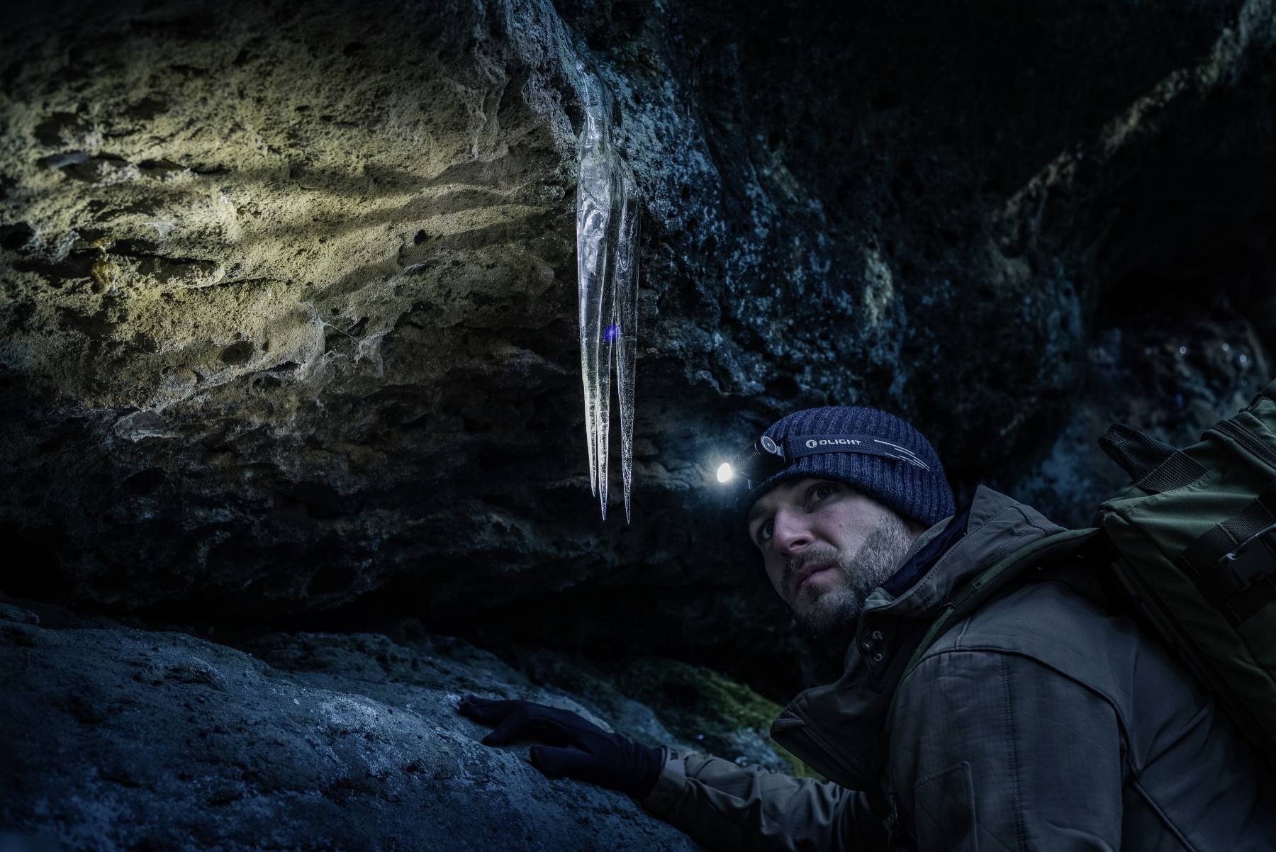 Eine Person in einem Wintermantel und mit einer Stirnlampe beobachtet Eiszapfen, die von der Decke einer dunklen Höhle hängen und durch das Licht der Stirnlampe beleuchtet werden.