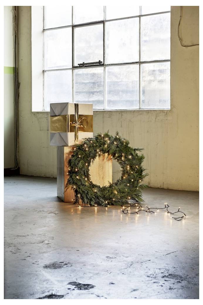 Ein Feiertagskranz, geschmückt mit Lichtern, lehnt sich gegen eine eingepackte Geschenkbox auf einem Betonboden in der Nähe eines großen Fensters und deutet auf Weihnachtsdekoration hin.