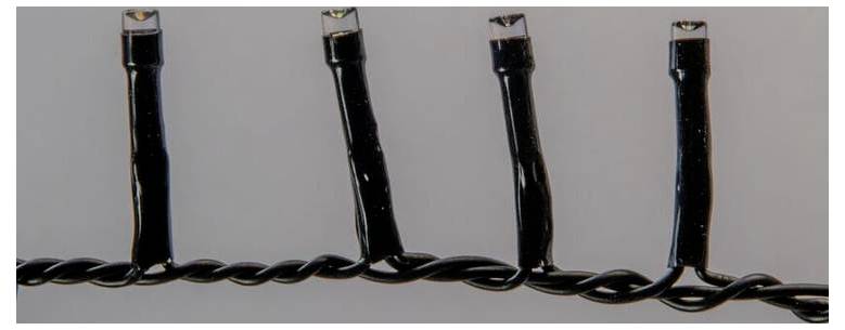 Eine Nahaufnahme mehrerer kleiner LED-Lichter auf einem schwarzen verdrehten Draht, möglicherweise Teil einer Reihe von Dekorationslichtern. Der Hintergrund ist grau.