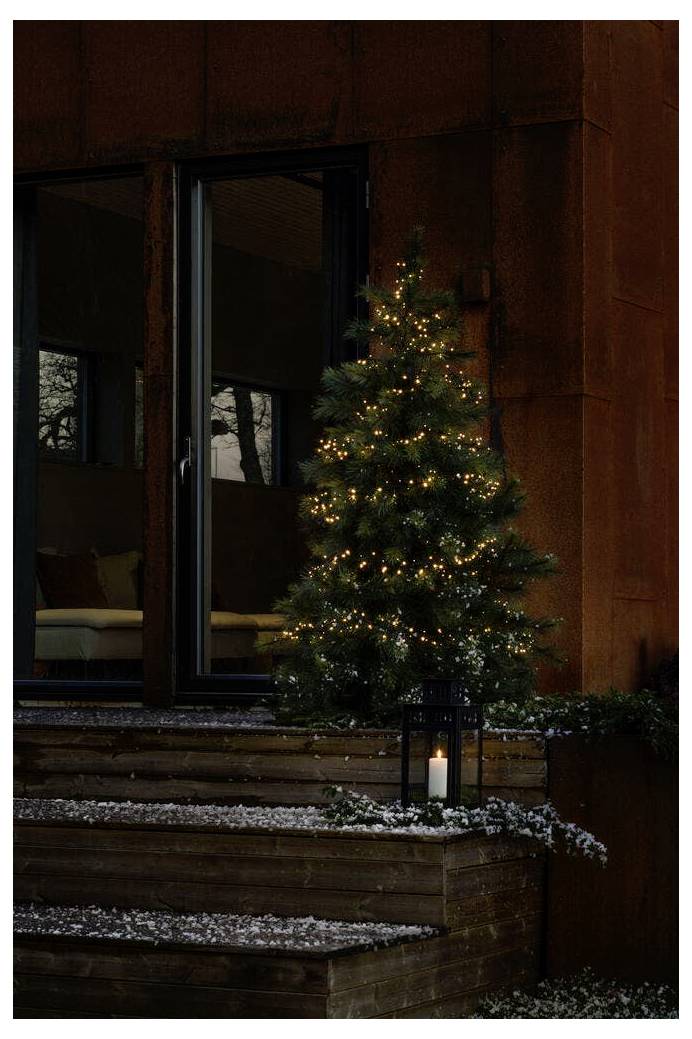Ein kleiner Weihnachtsbaum mit Lichtern steht neben einer Kerzenlaterne auf Holzstufen, daneben ein dunkles, modernes Haus mit Glastüren.