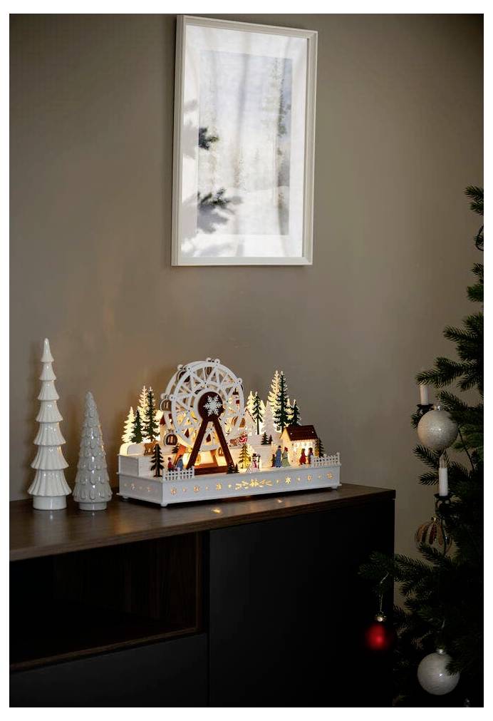 Dekorative Holzszene mit beleuchtetem Riesenrad, Bäumen und Häusern auf einem Schrank neben Weihnachtsbaumschmuck. Schatten auf Wandkunst.