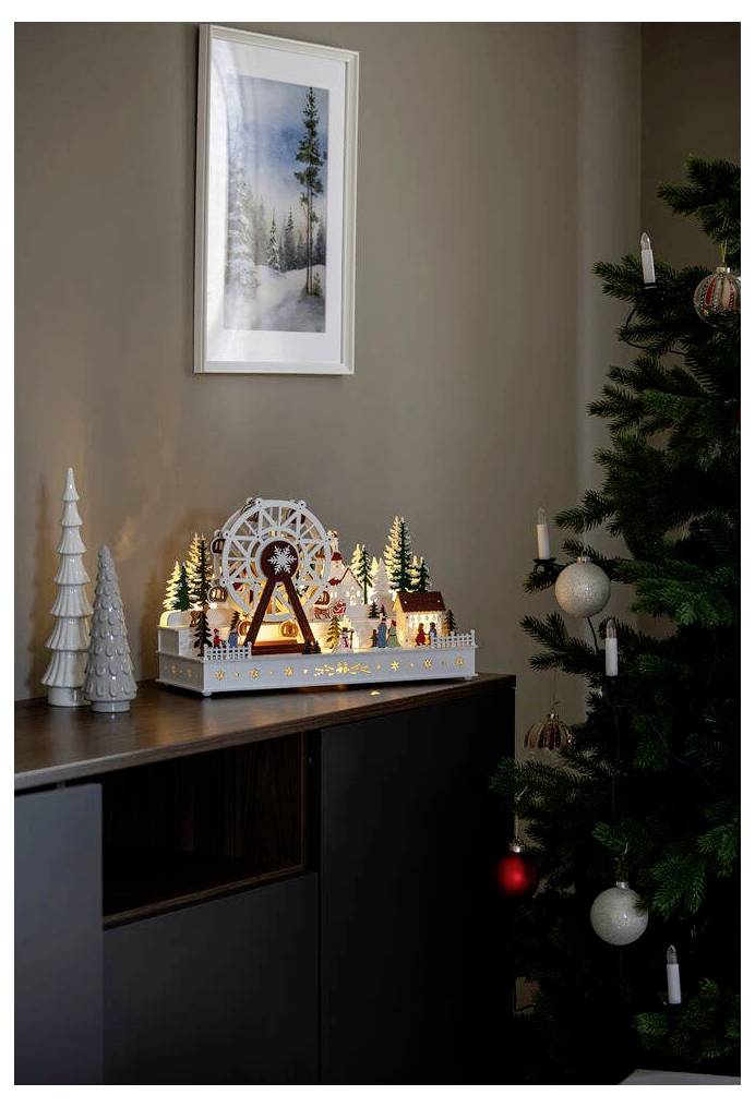 Eine gemütliche Weihnachtsszene mit einem geschmückten Baum und einer beleuchteten Weihnachtsdorf-Komposition auf einem Schrank, die ein Riesenrad und verschneite Bäume umfasst.
