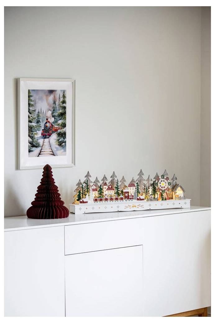 Ein dekorativer weißer Regal zeigt ein Papierweihnachtsdorf mit beleuchteten Häusern und Bäumen, unterhalb eines gerahmten Winterbildes mit einem Zug in verschneiten Wäldern.