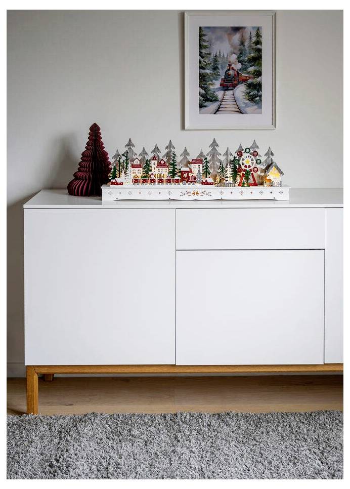 Ein weißer Schrank präsentiert ein festliches Modellgdorf mit bunten Gebäuden und Bäumen. Darüber hängt ein gerahmtes Winterbild mit einem Zug.