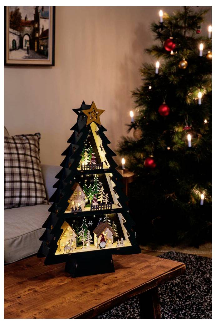 Eine Weihnachtsbaumdekoration aus Holz mit beleuchteten Ausschnitten von schneebedeckten Häusern und Bäumen steht auf einem Tisch in der Nähe eines beleuchteten Weihnachtsbaums.