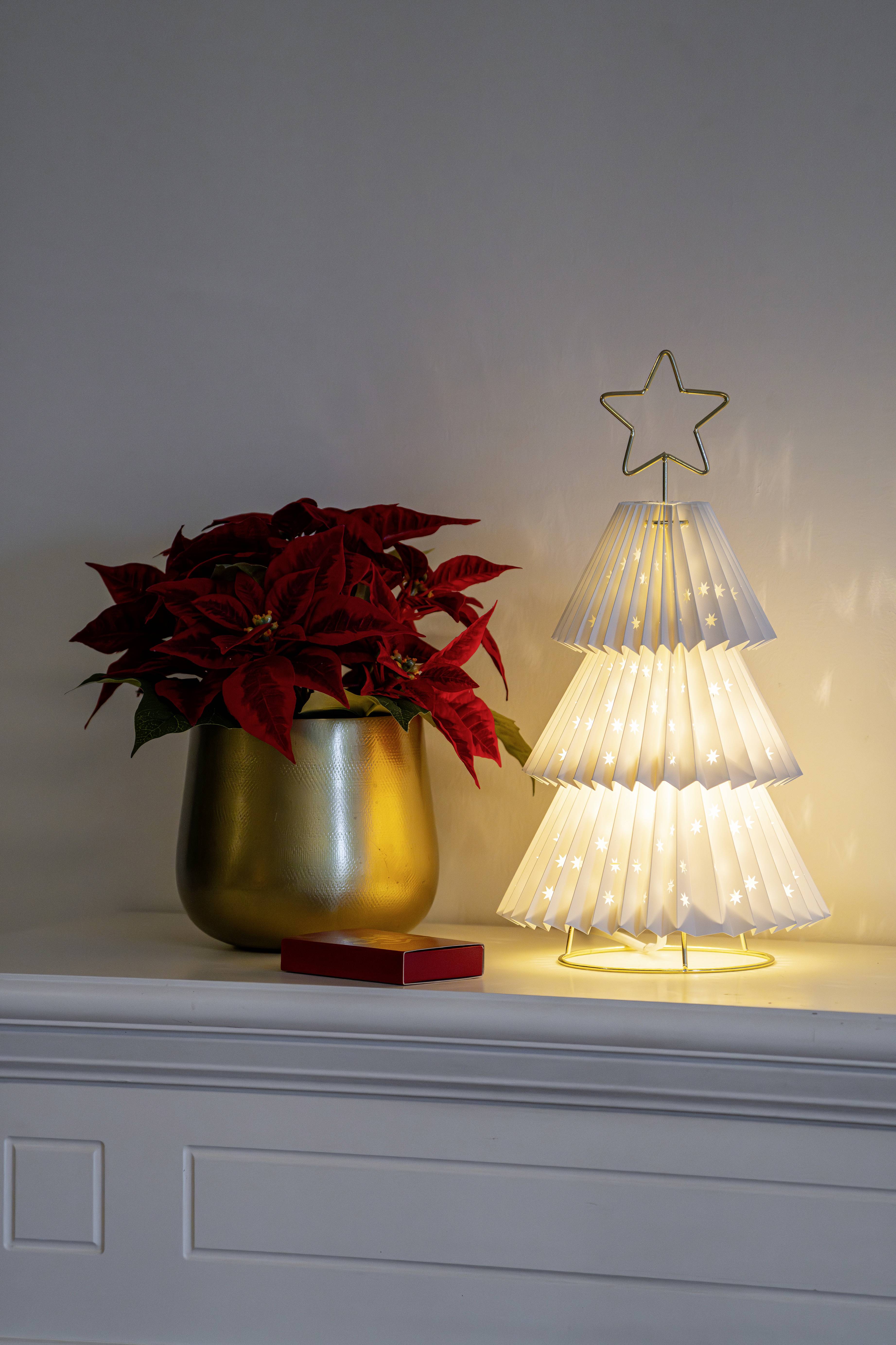 Dekorative weiße Papierlampe in Baumform mit Sternenspitze neben einer Weihnachtsstern-Pflanze in einem goldenen Topf, platziert auf einem weißen Kaminsims.