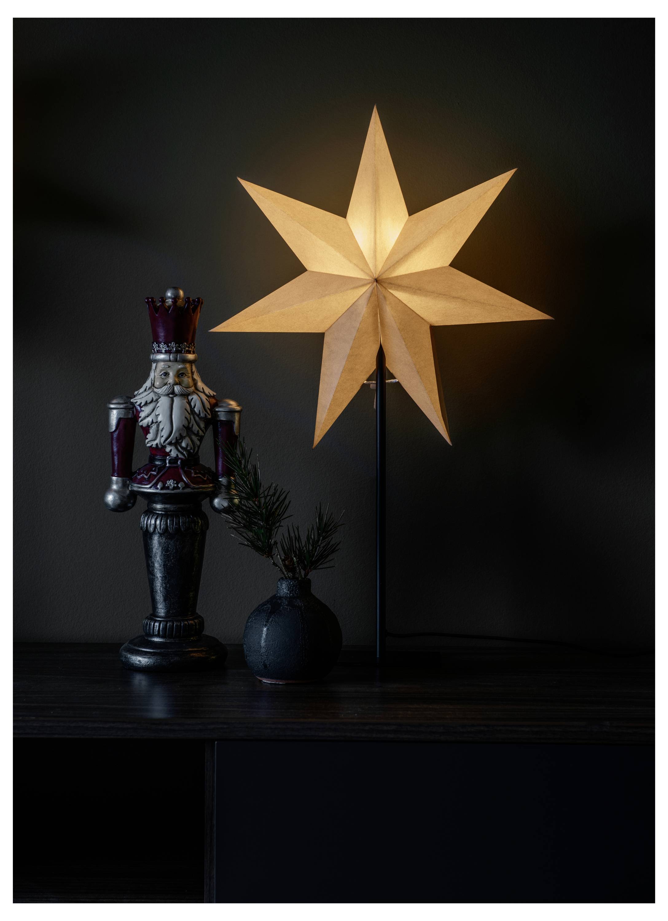 Eine leuchtende sternförmige Lampe auf einem Ständer neben einer dekorativen Nussknackerfigur und einer kleinen Vase mit Tannenzweigen auf einer dunklen Oberfläche.