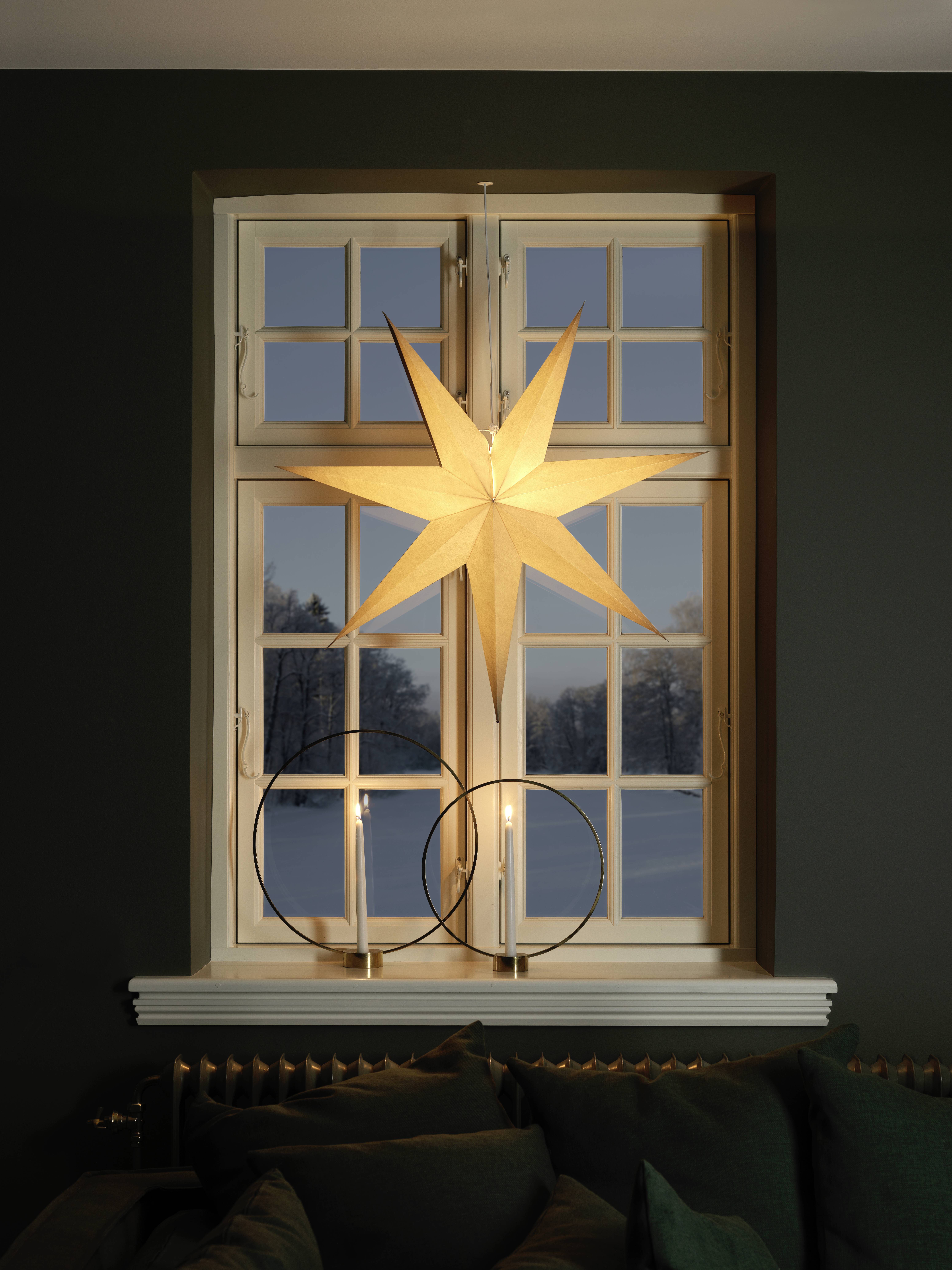 Ein großer beleuchteter Stern hängt in einem Fenster über zwei dekorativen Kerzenhaltern und verbreitet ein warmes Leuchten im dunklen Raum. Verschneite Landschaft draußen.
