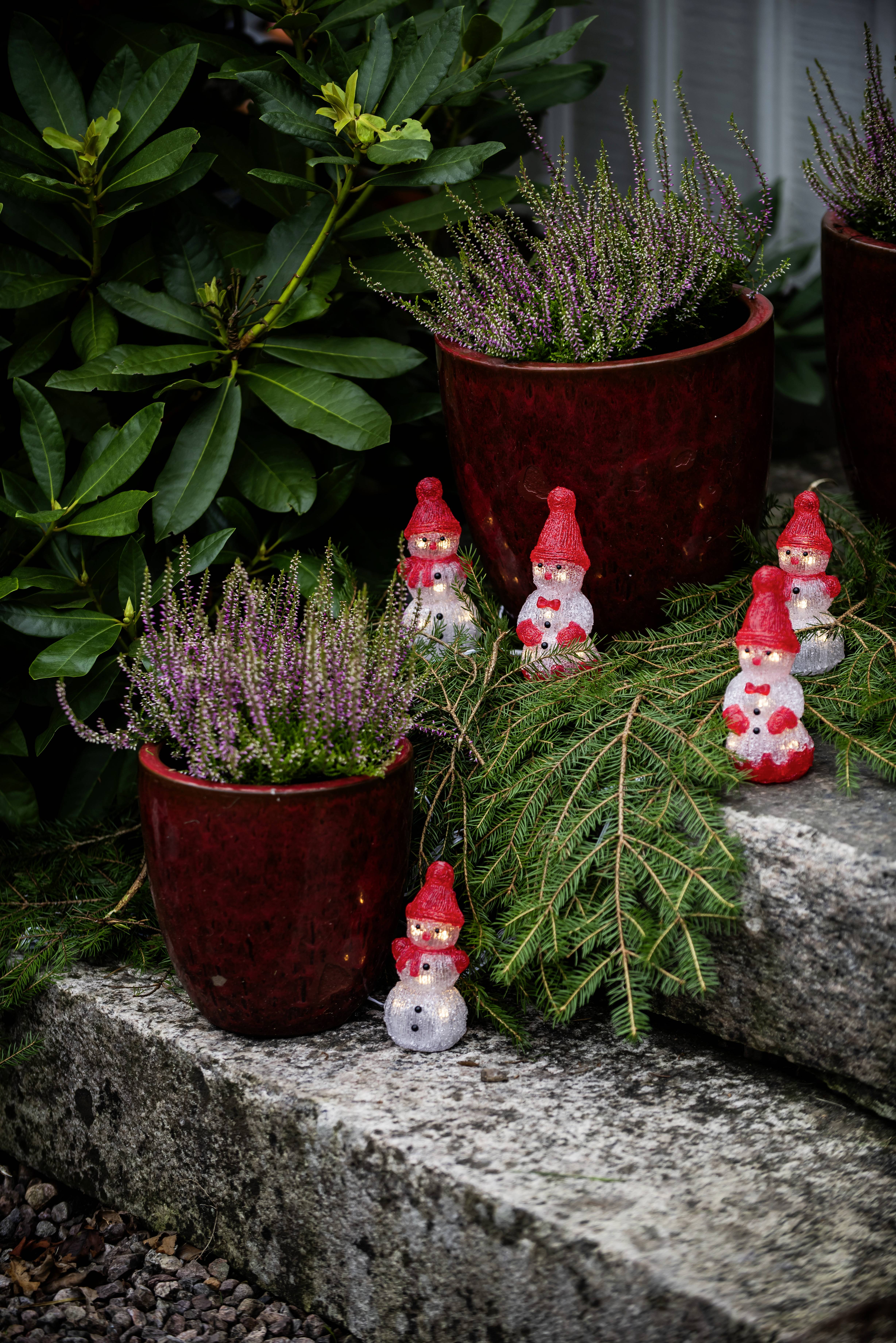Topfheide-Pflanzen in roten Töpfen sind auf Steinstufen angeordnet, umgeben von kleinen festlichen Gartenzwerg-Figuren und immergrünen Zweigen.