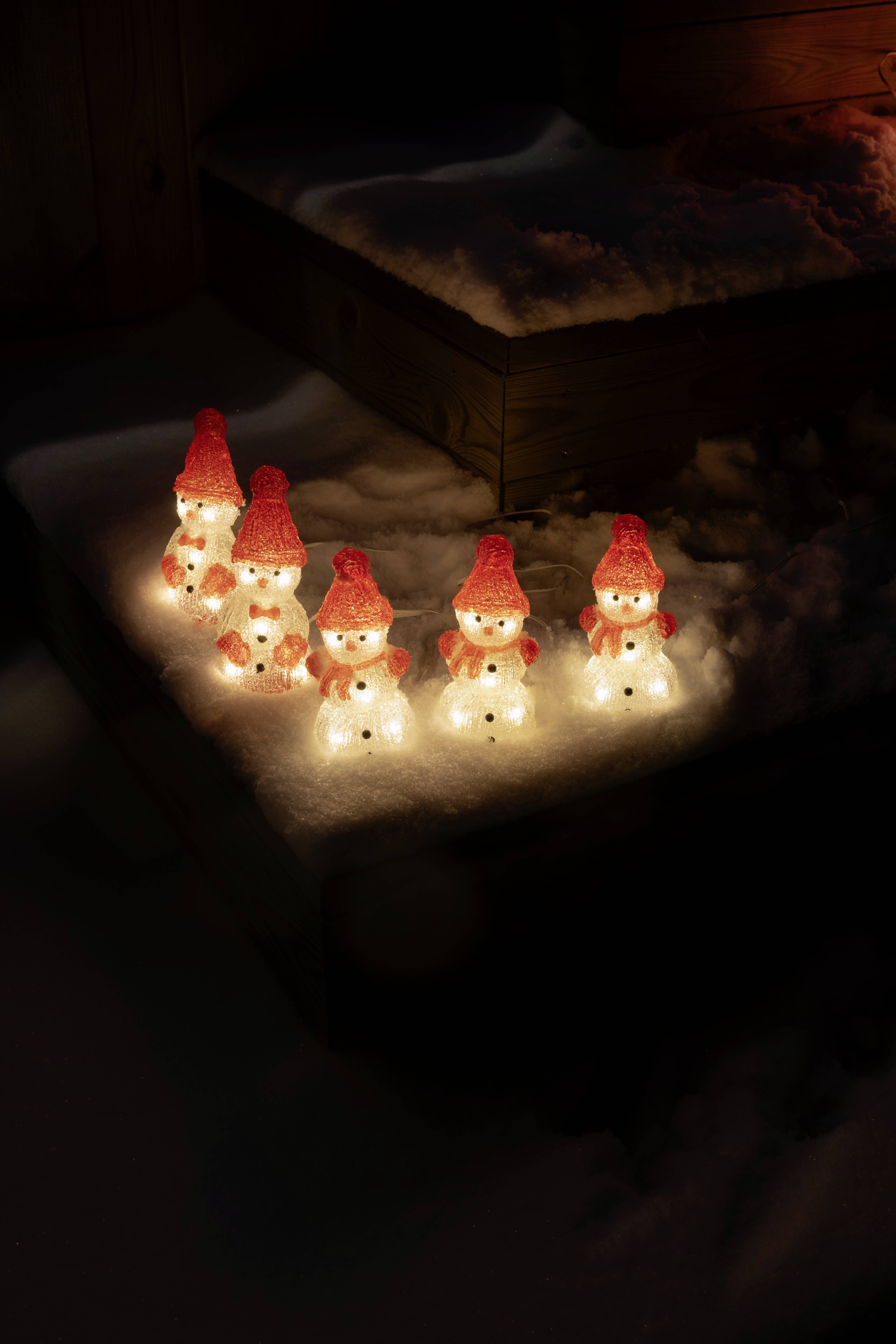 Eine Gruppe kleiner, beleuchteter Schneemann-Dekorationen mit roten Mützen, die auf verschneiten Holzstufen in der Nacht platziert sind und eine festliche Atmosphäre schaffen.