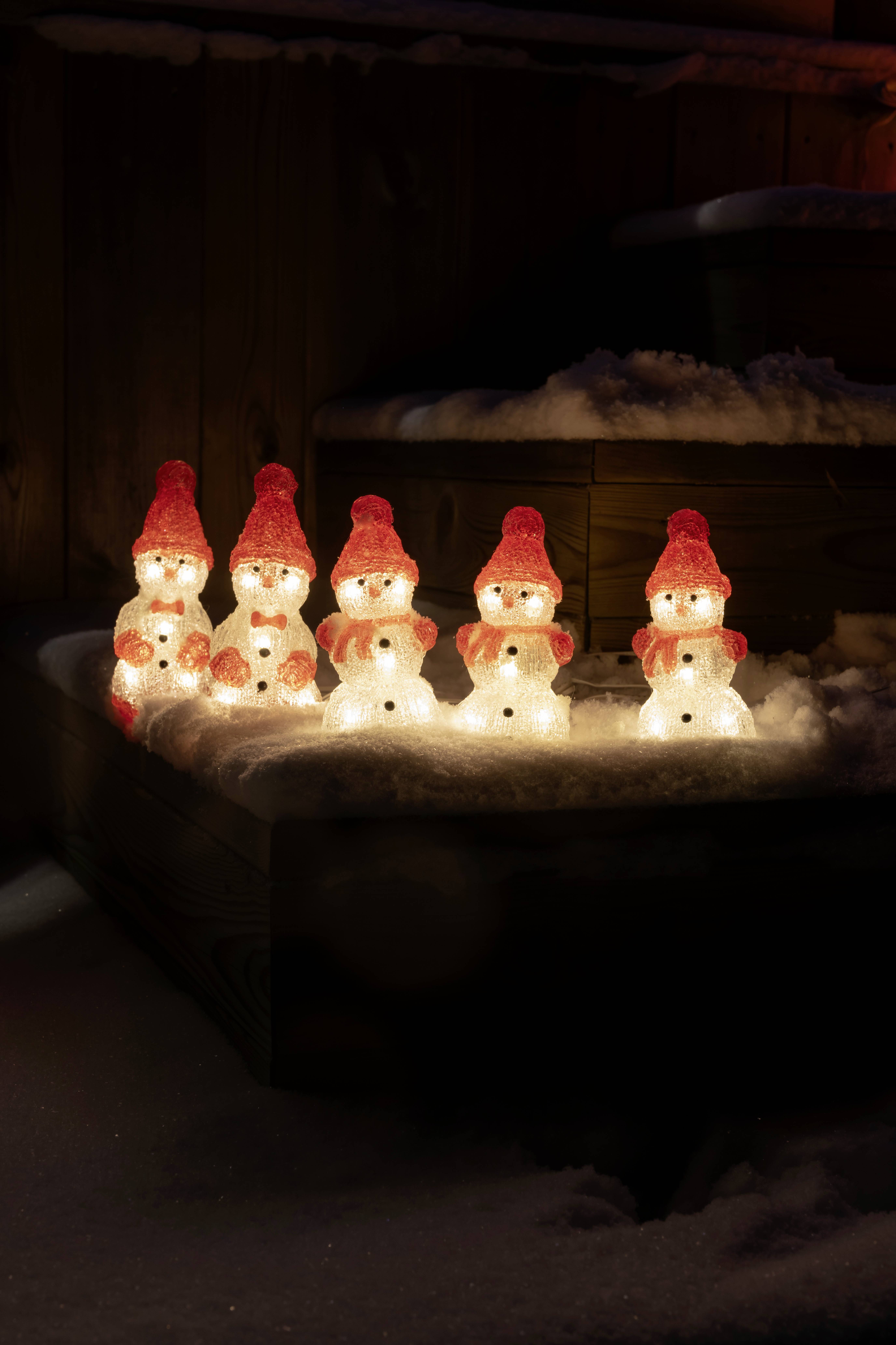 Fünf beleuchtete Schneemänner mit roten Hüten stehen in einer Reihe auf einer verschneiten Holzplattform in einer dämmrigen Umgebung.