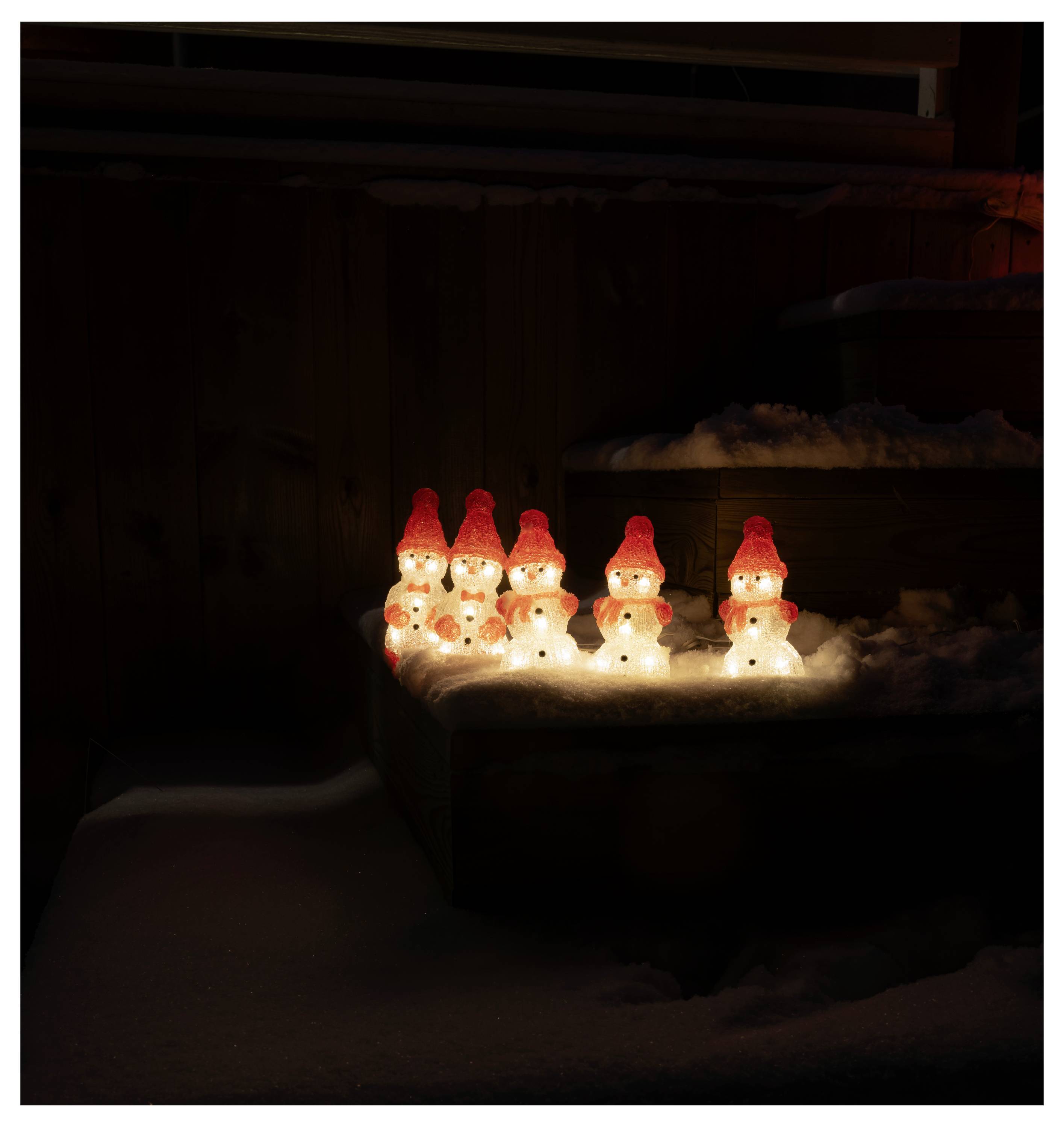 Eine Gruppe kleiner, beleuchteter Schneemännerfiguren mit roten Mützen, die nachts warm im Schnee leuchten und eine festliche Atmosphäre erzeugen.