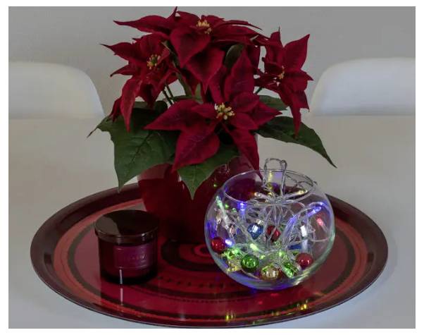 Eine rote Weihnachtsstern-Pflanze in einem Topf steht auf einem roten Tablett, begleitet von einer kleinen Kerze und einer Glaskugel, die mit festlichen Lichtern und Dekorationen gefüllt ist.