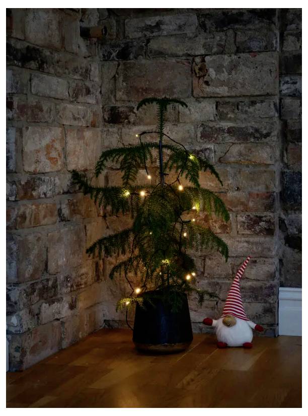 Ein kleiner Topfbaum mit Lichterketten steht in einer Ecke an einer Ziegelwand. Ein Gartenzwerg mit einer rot-gestreiften Mütze sitzt auf dem Boden daneben.