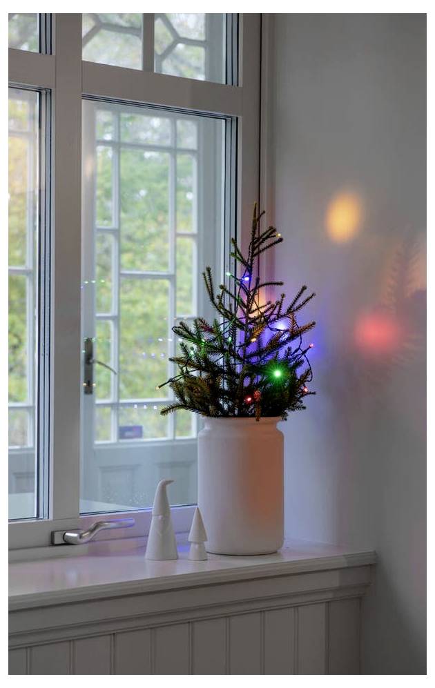 Ein kleiner, hell beleuchteter Weihnachtsbaum mit bunten Lichtern steht auf einem weißen Fenstersims neben einem Fenster und spiegelt Farben an der Wand wider.