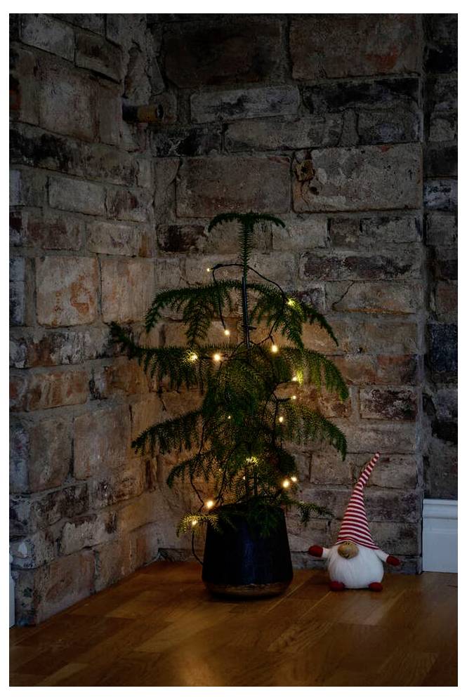 Ein kleiner, topfbepflanzter Immergrüner, der mit warmen gelben Lichtern geschmückt ist, steht in der Ecke eines Zimmers mit Ziegelwänden, und daneben befindet sich ein plüschiger Weihnachtsschmuck.