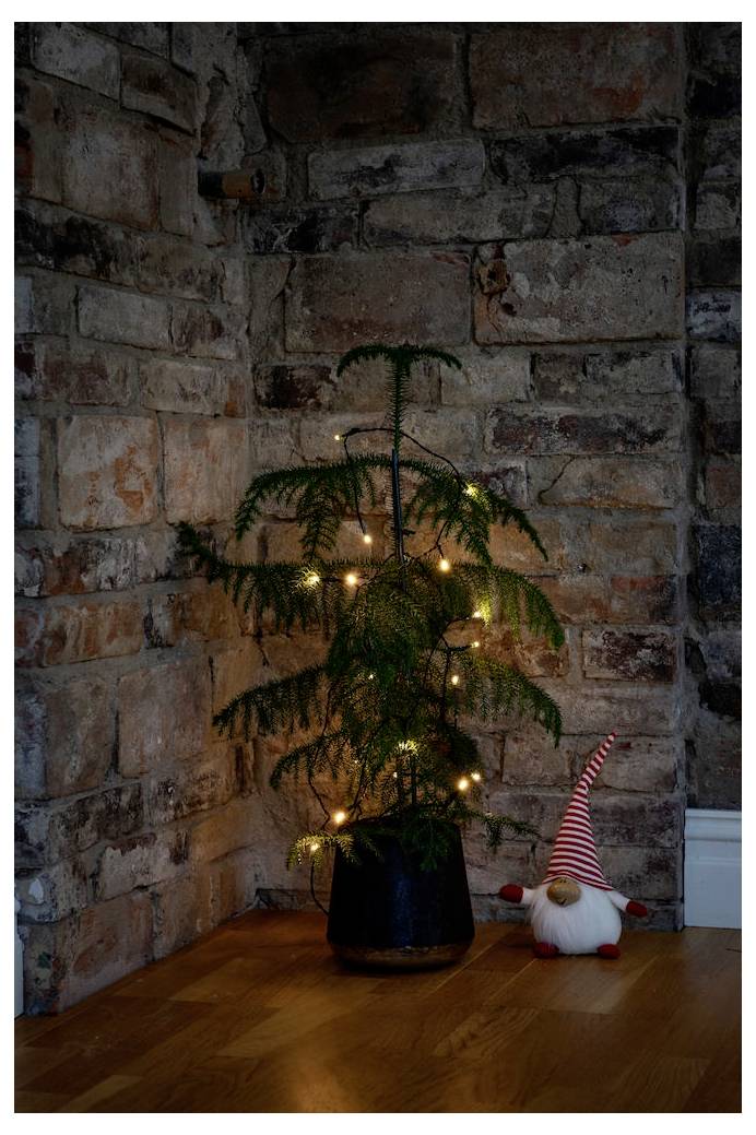 Ein kleiner Topfbaum mit Lichtern steht in einer Ecke an einer Ziegelwand. Eine festliche Gnom-Figur mit roter Mütze steht neben dem Topf.